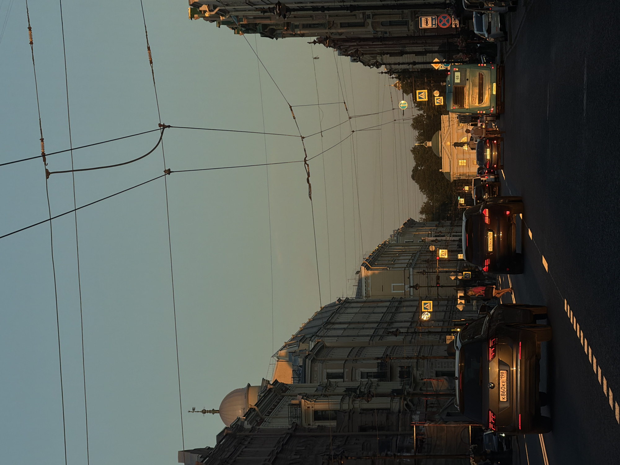 The image depicts a wide, straight street in Saint Petersburg, Russia, likely in the historic city center, as suggested by the classical architecture of the buildings lining both sides of the road. The time appears to be late afternoon or early evening, with a warm, golden light casting long shadows and illuminating the facades of the buildings. The sky is a clear, pale blue, transitioning to a soft yellow near the horizon.
Overhead, a complex network of trolleybus wires crisscrosses the sky, a common sight in Saint Petersburg. The street is moderately busy with vehicular traffic, including cars, a trolleybus (numbered 65), and possibly a bus. Several vehicles are captured in motion, with their headlights or taillights illuminated.
Pedestrians are visible on the sidewalks and crossing the street, though they are small in the frame. One woman in a bright pink top is walking on the sidewalk. The architecture features ornate facades, balconies, and domed structures, indicative of imperial Russian style. Trees are visible on the sides of the street, providing some greenery. Traffic signals and street signs are present, including pedestrian crossing signs. The road surface appears to be asphalt with white lane markings. The overall impression is of an active urban environment bathed in the soft, atmospheric light of a setting sun.