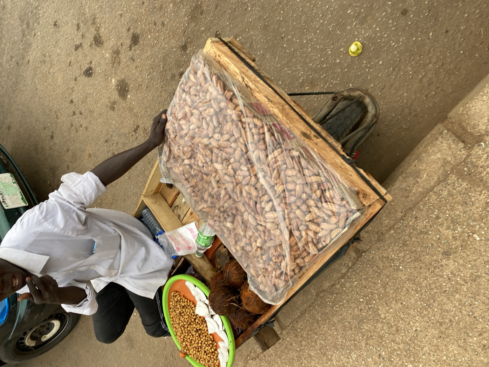 An overhead shot captures a street vendor in Bukuru, Nigeria, selling nuts and coconuts from a makeshift stall. The main subject is a dark-skinned individual, wearing a white striped long-sleeved shirt and black trousers, positioned beside a wooden cart or pallet. The vendor's right arm and hand are visible, holding or adjusting a large, clear plastic bag brimming with peanuts in their shells, which dominates the central part of the stall.
On the wooden stall, adjacent to the large peanut bag, are three fibrous brown coconuts. A vibrant orange and green plastic tray holds a heap of small, light brown roasted nuts or chickpeas, accompanied by some white pieces, possibly coconut flakes or another snack. A small green plastic bottle, clearly identifiable as "SPRITE," and a crumpled white plastic bag with some visible text, including "Y.Y. RABA," are also on the stall.
The setting is outdoors on a speckled grey and brown concrete surface, indicative of a street or market area. A car tire is partially visible to the left, and a light brown concrete wall or curb defines the right edge of the frame. A small yellow object, possibly a bottle cap, lies on the concrete ground in the upper right. The daylight illumination and the vendor's slight smile, visible in a cropped section, suggest a routine, daytime commercial activity.