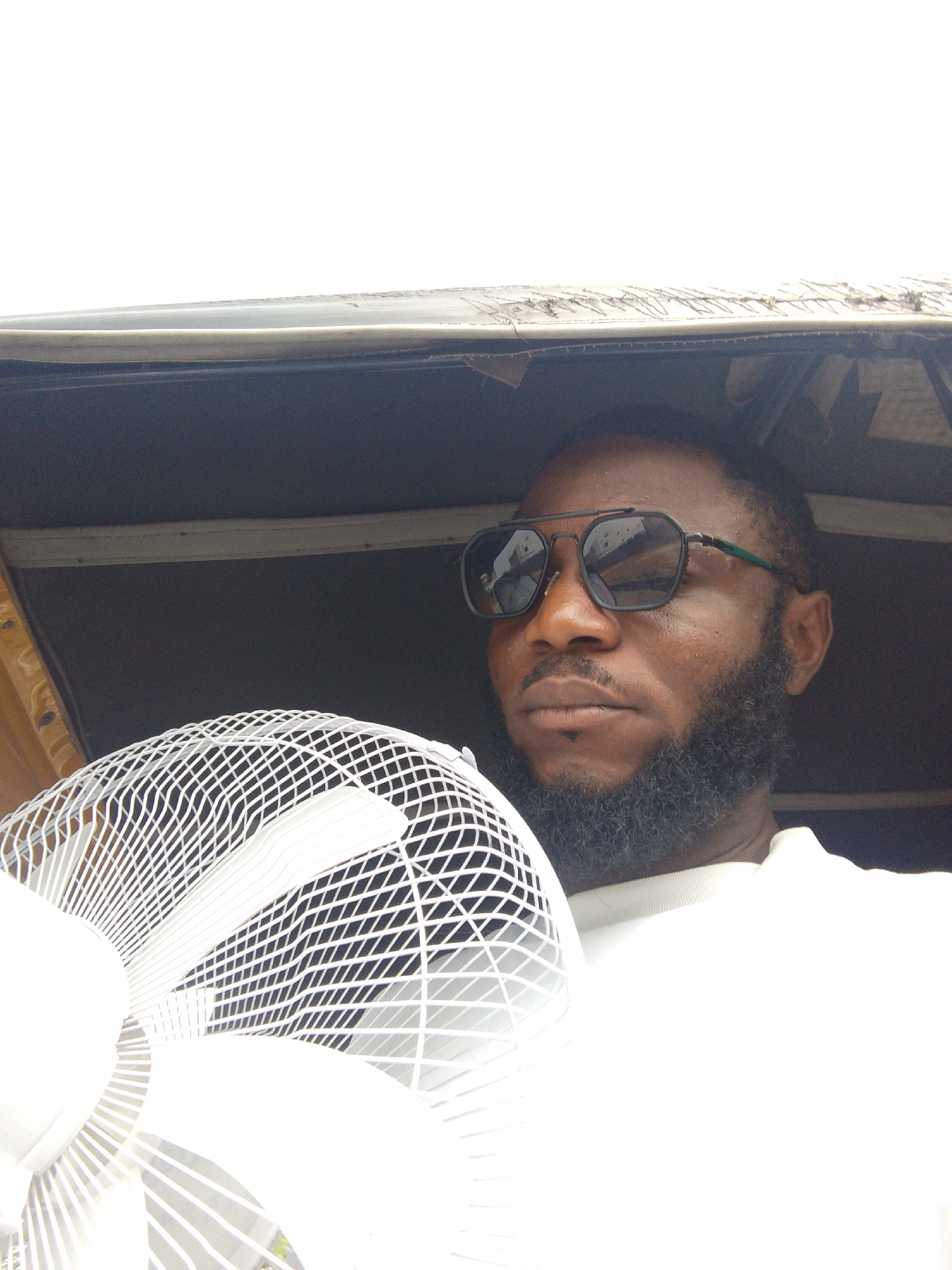 A medium-shot image captures a man with a dark complexion, a full beard, and sunglasses, holding a white electric fan. He is positioned within a three-wheeled vehicle (keke or auto-rickshaw) as the ceiling of the vehicle is visible. Based on the background details and the presence of the keke, the location is likely Lokoja, Nigeria. The man appears to be using the fan to cool himself.