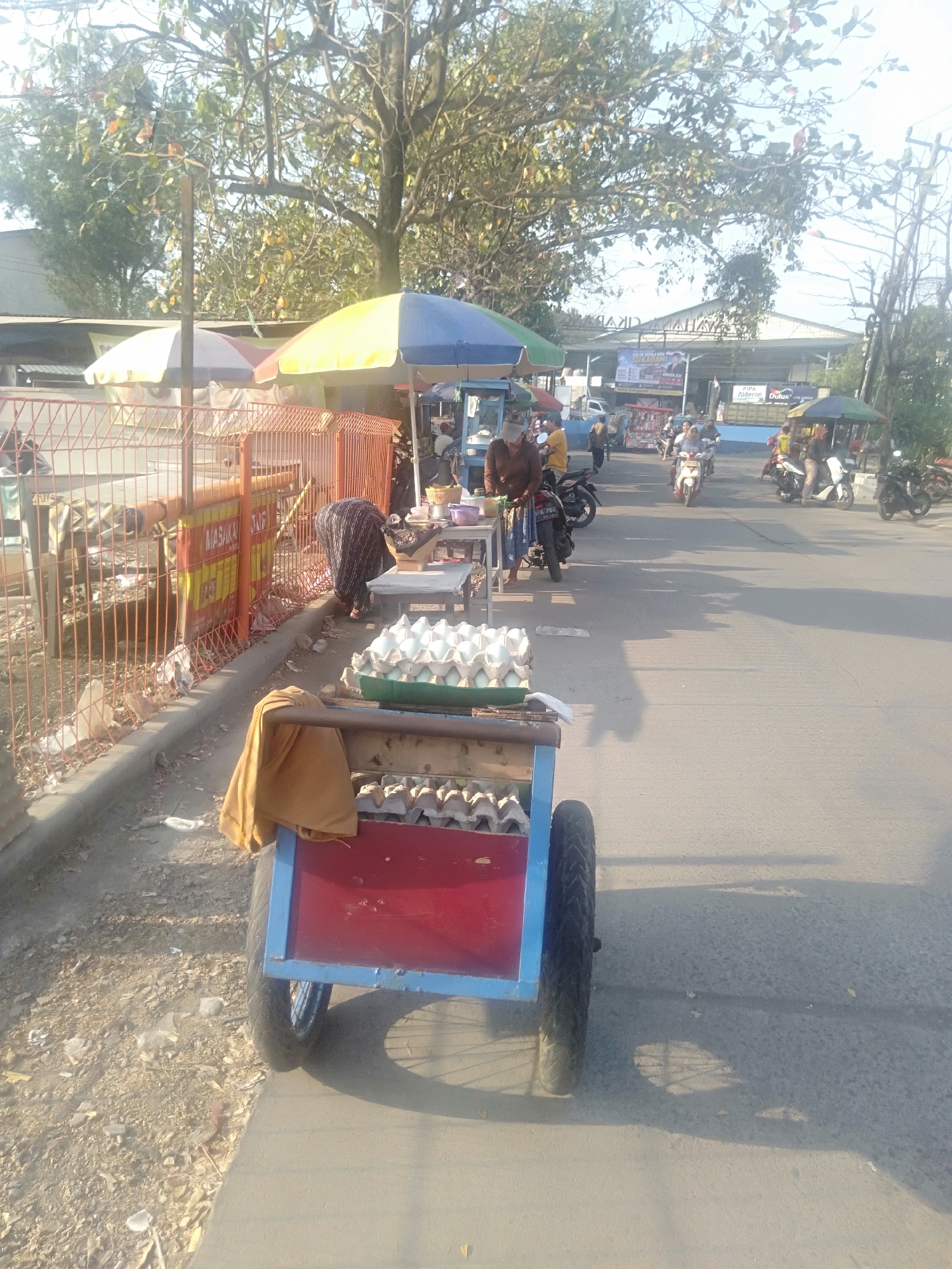 This is a medium-shot, eye-level, outdoor photograph taken on a sunny day in the city of Sukadami, Indonesia. The image captures a street scene with a market stall and passing vehicles. In the foreground, a red and blue cart is parked, loaded with several trays of light green eggs. A light brown cloth is draped over the side of the cart. Behind the cart, a woman in a patterned dress is bending over, tending to a stall. The stall has a table with various items, including a basket and bowls, and is partially covered by a multi-colored umbrella.
To the left of the stall, a red metal fence runs along the sidewalk, with a sign in yellow and red attached to it. A bare tree with sparse leaves stands behind the fence. Further down the street, several motorcycles are visible, some with riders. One motorcycle is parked near the woman's stall, and another is being ridden towards the viewer. In the background, more market stalls with umbrellas and awnings are set up, and a building with a sign is visible. The street is paved, and the ground on the side of the road is dirt and debris. The overall atmosphere suggests a lively street market environment.