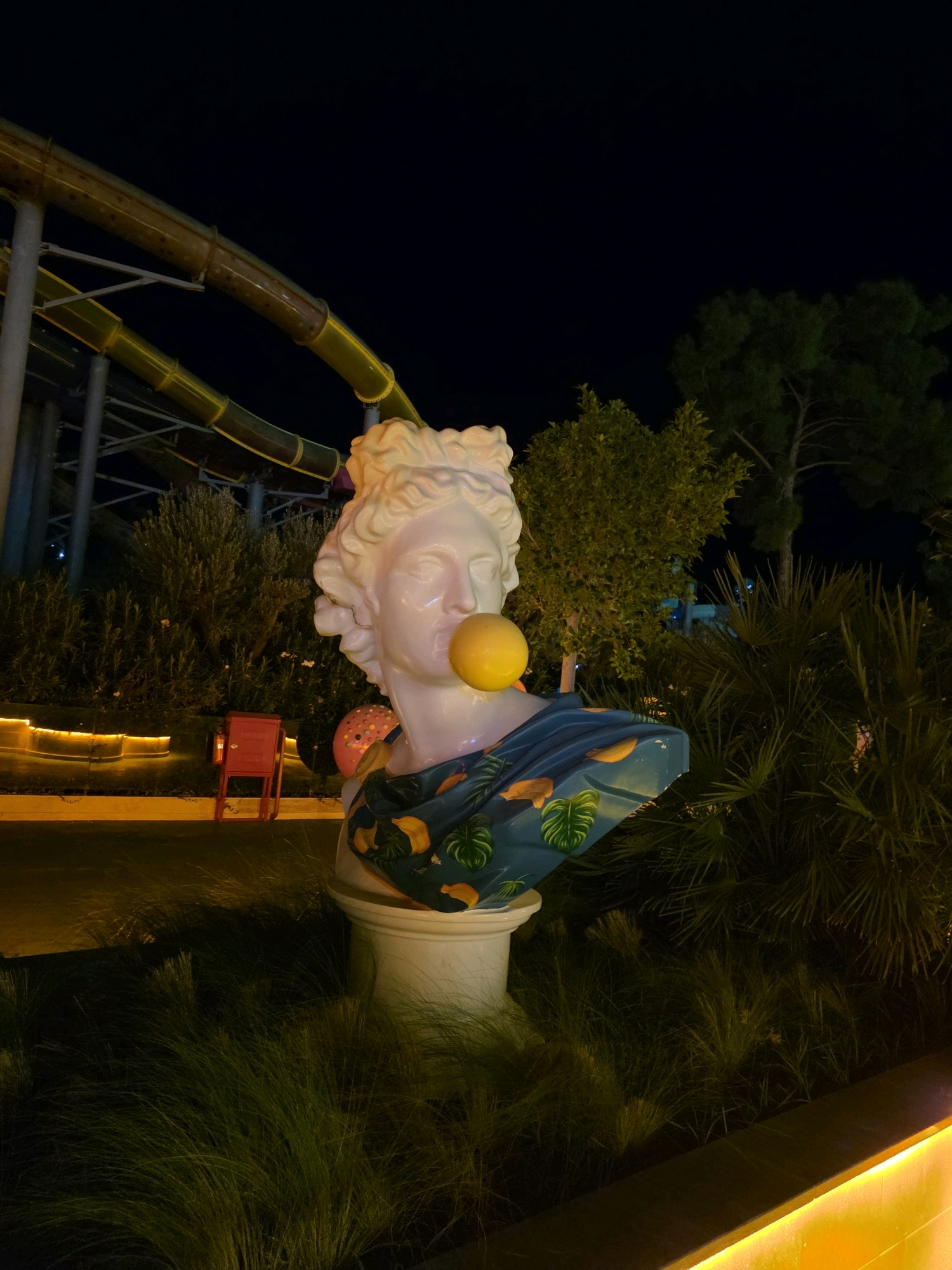 The image depicts a white classical bust statue of a male figure with curly hair, with a large yellow bubble protruding from its mouth. The statue is adorned with a draped fabric featuring a tropical print with bananas and palm leaves. It is mounted on a white pedestal and surrounded by lush green foliage. In the background, a section of a yellow water slide is visible, illuminated against a dark night sky. To the left of the statue, a red metal object, possibly a trash can or fire extinguisher stand, is situated. The scene appears to be an outdoor setting at night, likely a resort or entertainment venue. The context of Bodrum, Turkey, suggests this might be a decorative element in a public space or a resort area, typical of tourist destinations.