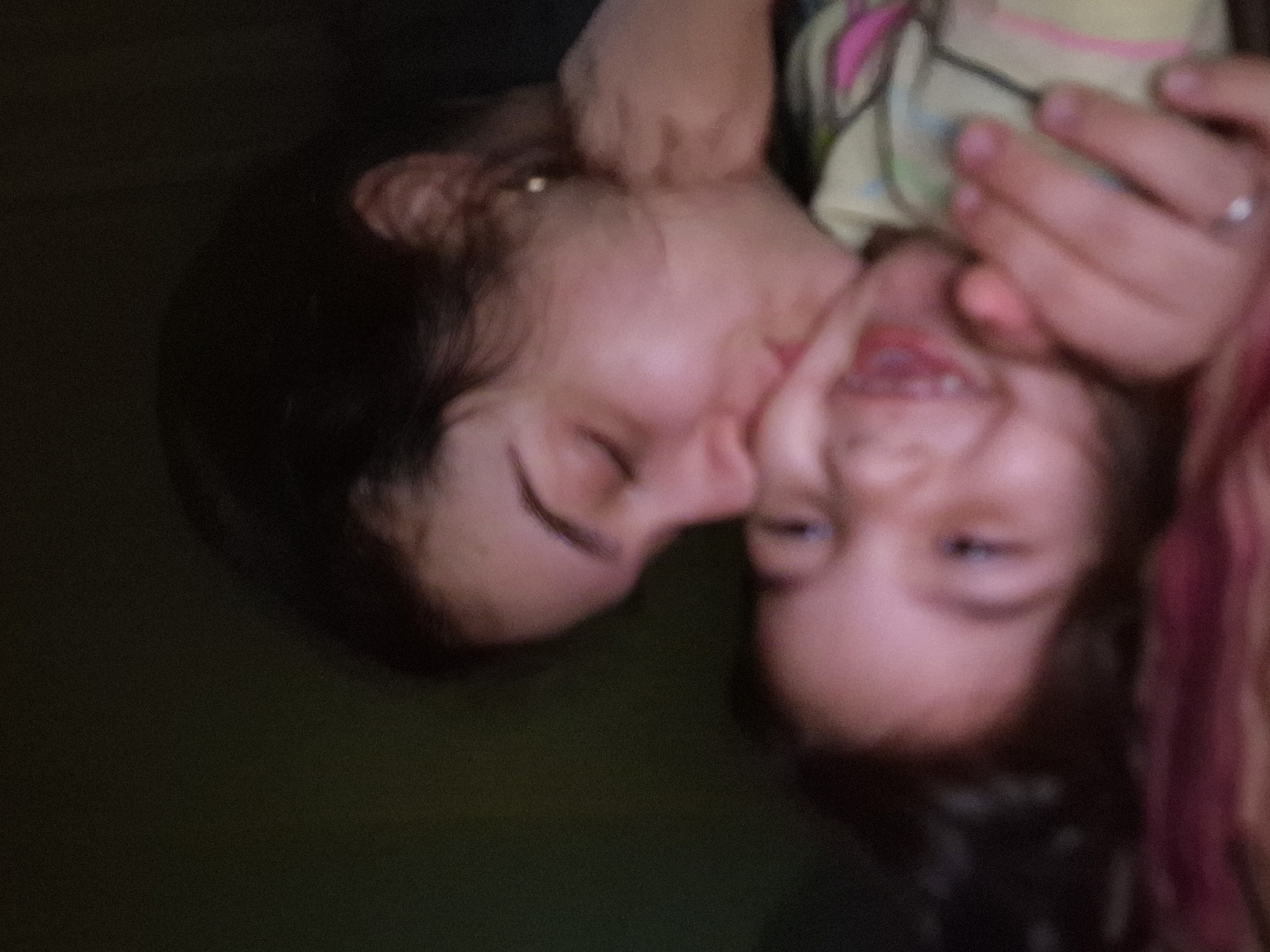An out-of-focus, grainy image shows two individuals, a child and an adult female. The adult female, positioned on the left, has dark hair, and her eyes are closed. Her skin tone is light. To her right is a child smiling with visible teeth. The background is a dark green. It is not possible to identify any specific location in Byala Slatina, Bulgaria from this image.