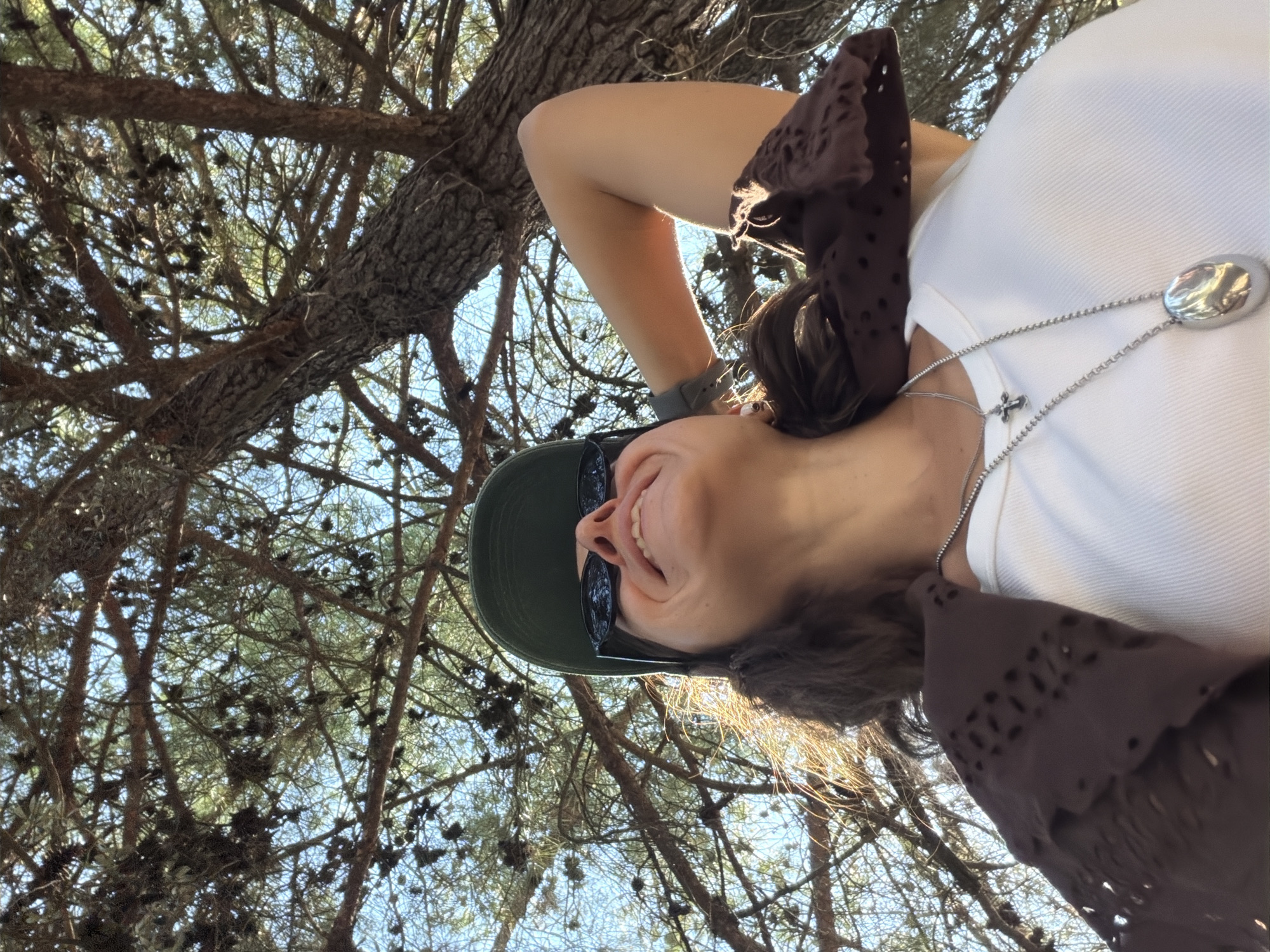 A low-angle shot captures a woman from the chest up, looking upwards towards the sun and foliage of a tree. She is wearing a white tank top, a brown distressed mesh cardigan, a green baseball cap, and black cat-eye sunglasses. She has her right arm raised behind her head, with her elbow bent. A silver necklace with a cross charm and an oval pendant hangs around her neck. Her skin is illuminated by the sun, creating a warm glow. The background consists of the intricate branches and needles of a pine tree against a clear blue sky. The scene appears to be outdoors, likely in a park or natural setting within Budva, Montenegro, suggested by the presence of Mediterranean flora and bright sunlight.