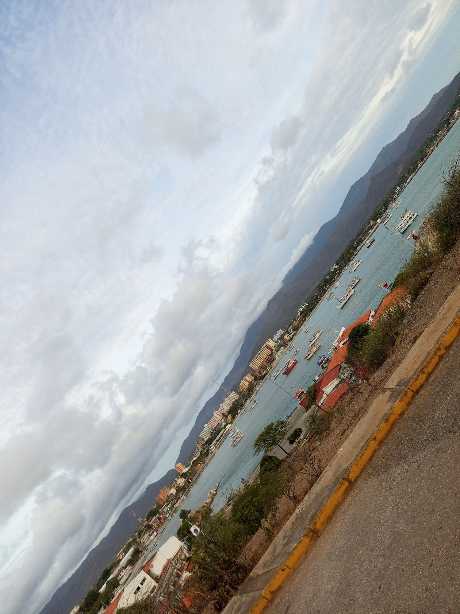 The image shows a cityscape with a bay and mountainous backdrop in Juan Griego, Venezuela. The photo is taken from an elevated position, capturing boats in the bay, buildings with orange roofs, and mountains in the distance. A cloudy sky is visible overhead. A sidewalk with a yellow curb borders the lower right of the frame, alongside a section of road.