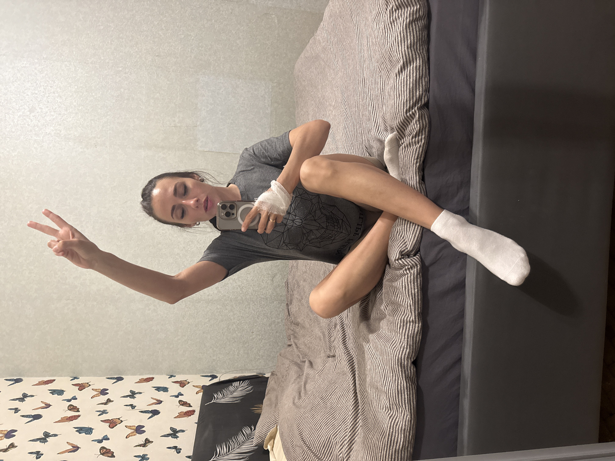 A young adult female, likely in her 20s or early 30s, is depicted in a full-length mirror selfie within a bedroom in Saint Petersburg, Russia. She is seated cross-legged on a bed, facing the mirror with her body slightly angled to the viewer's left. Her dark hair is pulled back, and she wears a grey short-sleeved t-shirt with a dark geometric design and white socks on both feet. Her left hand, positioned on her lap, is wrapped in a white bandage or gauze. She holds a silver-colored smartphone, equipped with three rear cameras and a visible MagSafe ring, in her right hand, using it to capture the image. Her right arm is raised, forming a "peace" sign with her index and middle fingers. Her eyes are partially closed or looking downwards towards the phone screen. A gold-colored ring is visible on her left ring finger.
The bed features a dark grey frame and fitted sheet, topped with a duvet cover displaying horizontal stripes in alternating light and dark brown tones. A dark grey pillow with a white feather pattern is visible on the left side of the bed. The wall behind the bed on the left features white wallpaper adorned with numerous colorful butterfly motifs (blue, orange, brown). The wall behind the bed on the right, and extending across the majority of the background, appears to be a light-colored, subtly textured mirrored surface, reflecting elements of the room and creating the visual context for the selfie.
