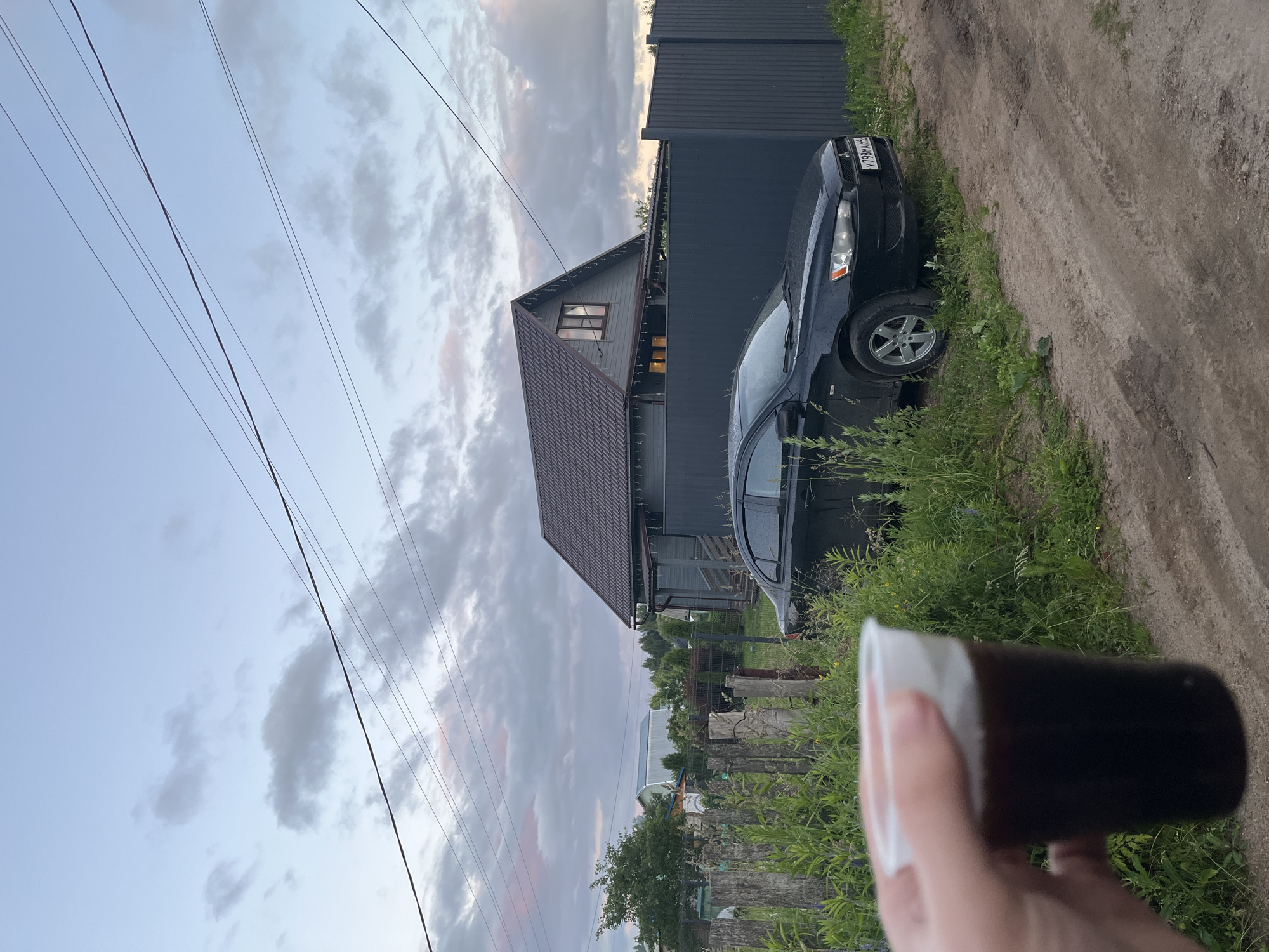 A hand holds a clear plastic cup containing a dark liquid and white foam. In the background, a gray, two-story house with a brown roof is visible. A dark-colored sedan is parked in front of the house. The scene is set in a rural area with a dirt road, overgrown grass, and a wooden fence. Overhead, power lines crisscross the sky, which is filled with clouds. The license plate of the car reads "Y798MA 56". The location is identified as Klobyushnevo, Russia.