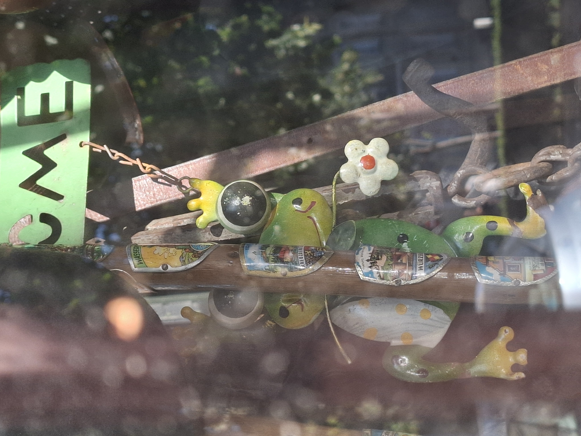 A reflection in a window obscures a vertical wooden post decorated with several attached decorative shield-shaped metal ornaments, each depicting a miniature scene. Attached to the post is a string of three frog-shaped figures, painted green and yellow. The top frog wears a yellow crown and has a black, round eye. The middle frog is also green and yellow with a black eye and a red mouth. The bottom frog has a similar design with a green body and yellow extremities. A white flower with a red center hangs from a chain attached to the middle frog. Above the frogs, a green metal sign with cut-out letters reads "COME". A rusty metal chain hangs down from the sign and connects to the top frog. The background is blurred, showing trees and foliage. The scene is likely outdoors, viewed from inside through a window.