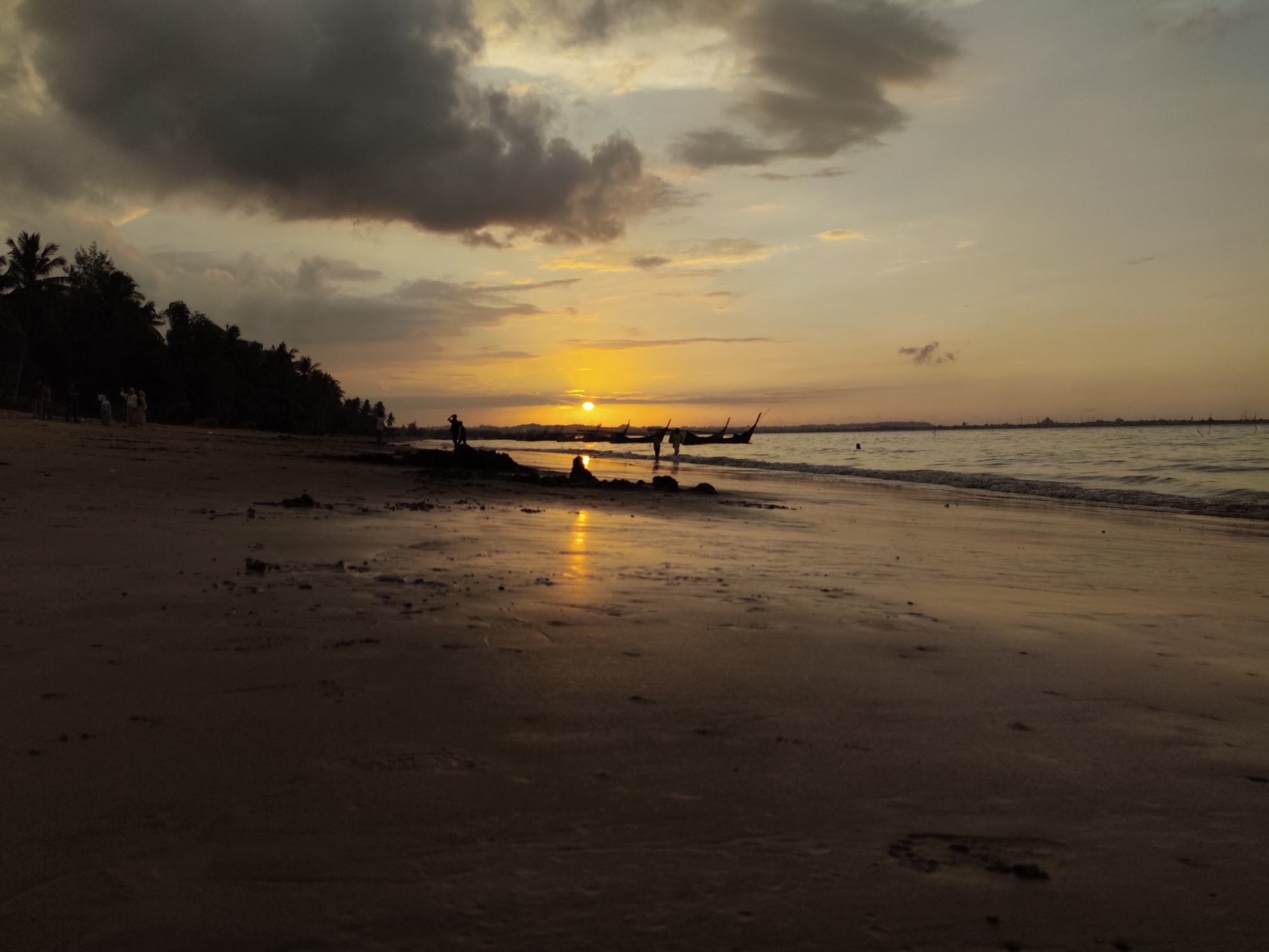 The image depicts a sunset scene at a beach, likely in Lhokseumawe, Indonesia, given the typical coastal landscape. Silhouetted against the golden hues of the setting sun are several people walking along the shoreline, their forms indistinct. A line of traditional fishing boats, also in silhouette, are moored or anchored in the calm water. The beach itself is sandy, with evidence of water receding from the tide, creating reflective patches of wet sand. In the background, a dense line of trees, likely palm trees, marks the edge of the land. The sky is a blend of warm oranges and yellows near the horizon, transitioning to darker, more dramatic gray clouds higher up, indicating a dynamic weather condition during the sunset. The overall impression is one of tranquility and natural beauty, with human activity subtly integrated into the environment.