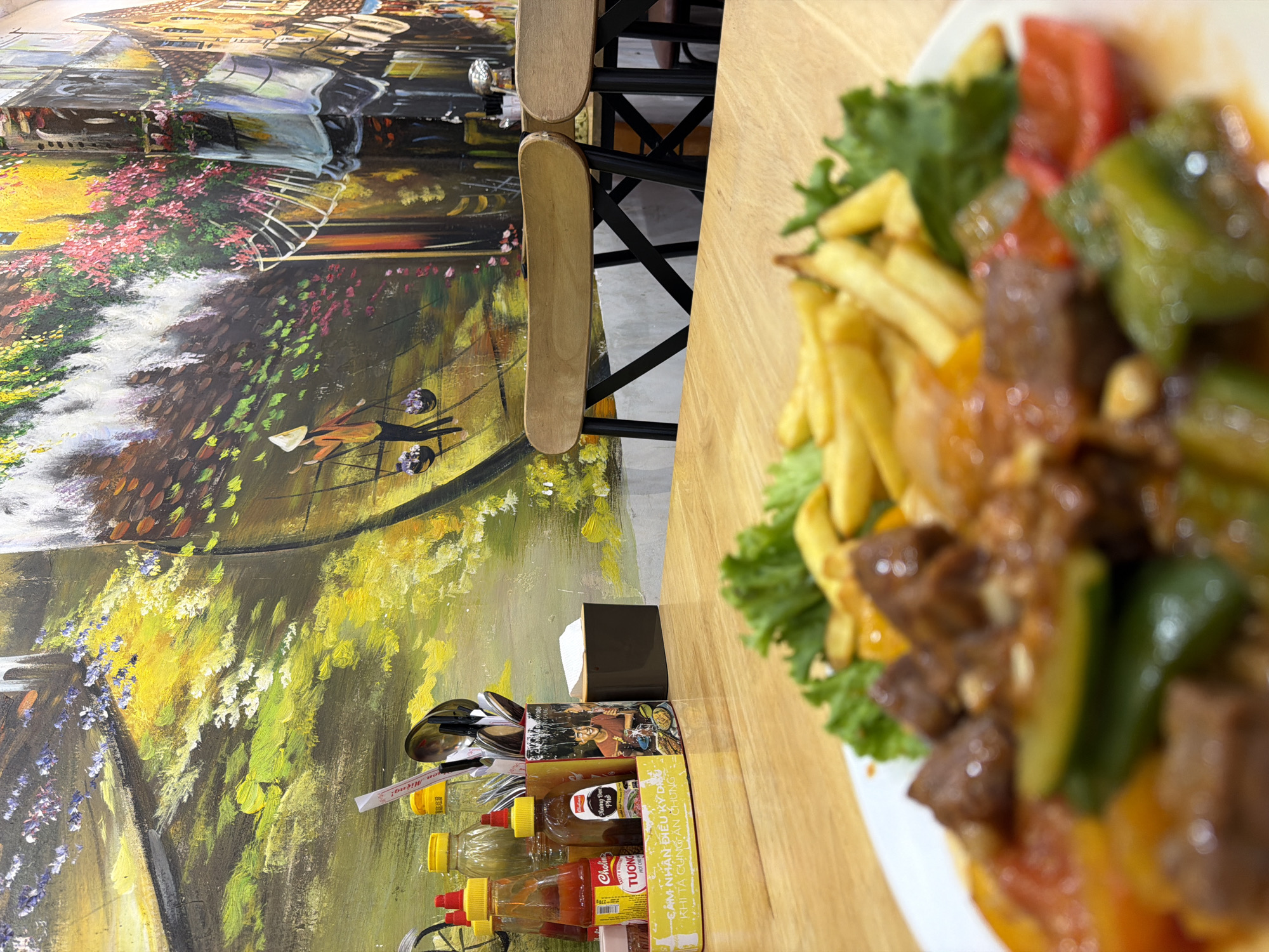 Here is a detailed description of the image:
**Setting:**
The photograph appears to have been taken in a restaurant or dining area. A large wall painting dominates the background, depicting a European-style cityscape with a bridge, water, and buildings. The painting exhibits various colors, including yellows, greens, browns, and blues.
**Foreground:**
The primary subject is a plate of food placed on a wooden table. The dish includes what appears to be fried potatoes (fries), lettuce, and a stir-fried mixture of meat cubes and colorful bell peppers (red, green, and yellow). There is also a condiment organizer with sauces, utensils, and napkins.
Nearby, there is a wooden chair
**Objects:**
* Food: Plate of beef with bell pepper and fries.
* Table: Wooden table.
* Chair: Wooden chair with a black metal frame.
* Condiments: Various sauces and utensils in a tray.
* Napkins: Black container holding napkins.
* Wall Art: Large painting behind the table.
**Location Information (Based on OCR Text):**
The text “Phở” suggests a Vietnamese setting. The text, "CẢM NHẬN ĐIỀU KỲ DIỆU KHI TÀ CÙNG ĂN CHƯNG Miệng!", on a condiment organizer suggests a location in Vietnam. The text "Nha Trang Ward" further specifies that the location of the photograph is in a ward of the city of Nha Trang.
**Overall Impression:**
The scene is likely a restaurant setting in Nha Trang, Vietnam, focusing on a meal being served. The wall art adds a distinct ambiance to the dining area.