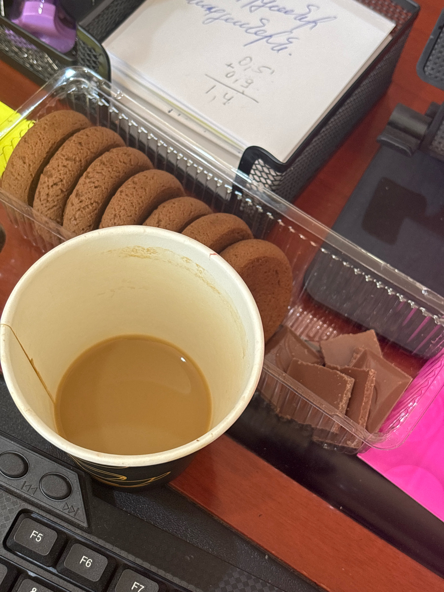 A close-up overhead shot shows a cup of coffee and a package of cookies on a desk. The coffee is in a white paper cup with gold detailing. The cookies, which are round and brown, are in a clear plastic container. Beside the cookies is a small container of chocolate pieces. A notebook with handwritten notes and numbers is visible in the background, next to a black mesh desk organizer. The desk appears to be made of dark wood. The image is taken from a high angle, looking down at the objects on the desk. There is no indication of the location in Ijevan, Armenia.