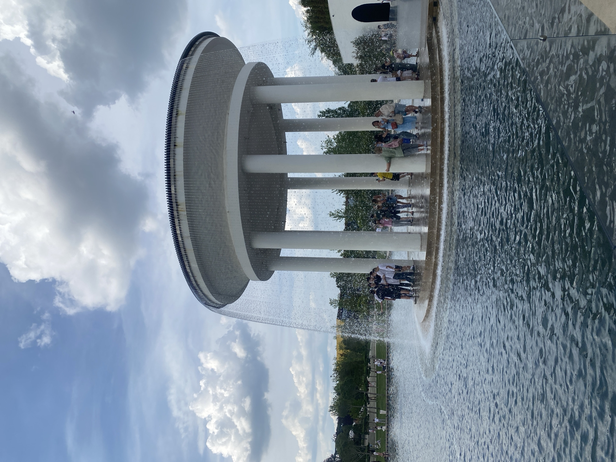 This image depicts a contemporary circular "infinity fountain" structure situated within a large artificial pond, likely in Galitsky Park (Krasnodar Park) in Krasnodar, Russia. The fountain consists of a broad, flat, white circular canopy supported by multiple white cylindrical columns, from which water cascades downwards in a continuous sheet and individual streams, creating a curtain effect.
Numerous individuals are gathered around and within the fountain structure. A group of approximately seven young males is positioned together on the circular platform directly beneath the falling water on the left side, facing towards the right. Other people, including adults and children, are distributed around the platform, some standing under the falling water, others observing from the edge. They are dressed in casual summer attire.
The fountain is surrounded by a body of calm, dark blue-green water, exhibiting ripples on its surface. Beyond the pond, a manicured park environment is visible, featuring green lawns, various trees, and distant park visitors. A modern white building with a dark arched entrance is partially visible on the far right. The sky above is partly cloudy, with large white cumulus clouds against a lighter blue sky, suggesting bright daylight conditions. A faint, dark object, possibly a bird or drone, is visible in the sky above the fountain in one of the crop images.