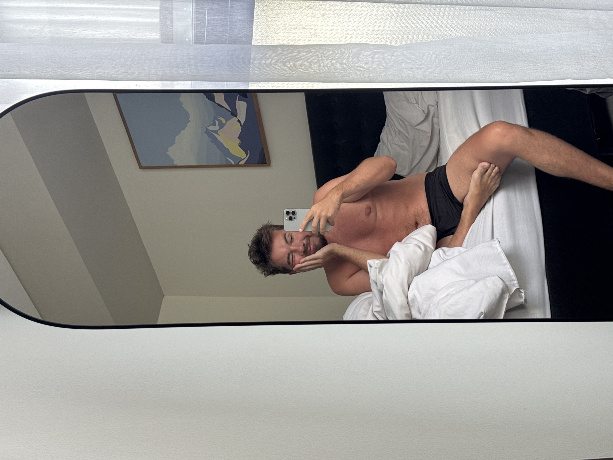A mirror on a white wall reflects a man in black boxer shorts lying on a white-sheeted bed. He is holding a smartphone and posing with his hand on his cheek. A framed painting hangs on the wall behind the bed. The top of the mirror reflects a partially visible window with closed blinds. The setting appears to be a room in North Kuta, Indonesia.
