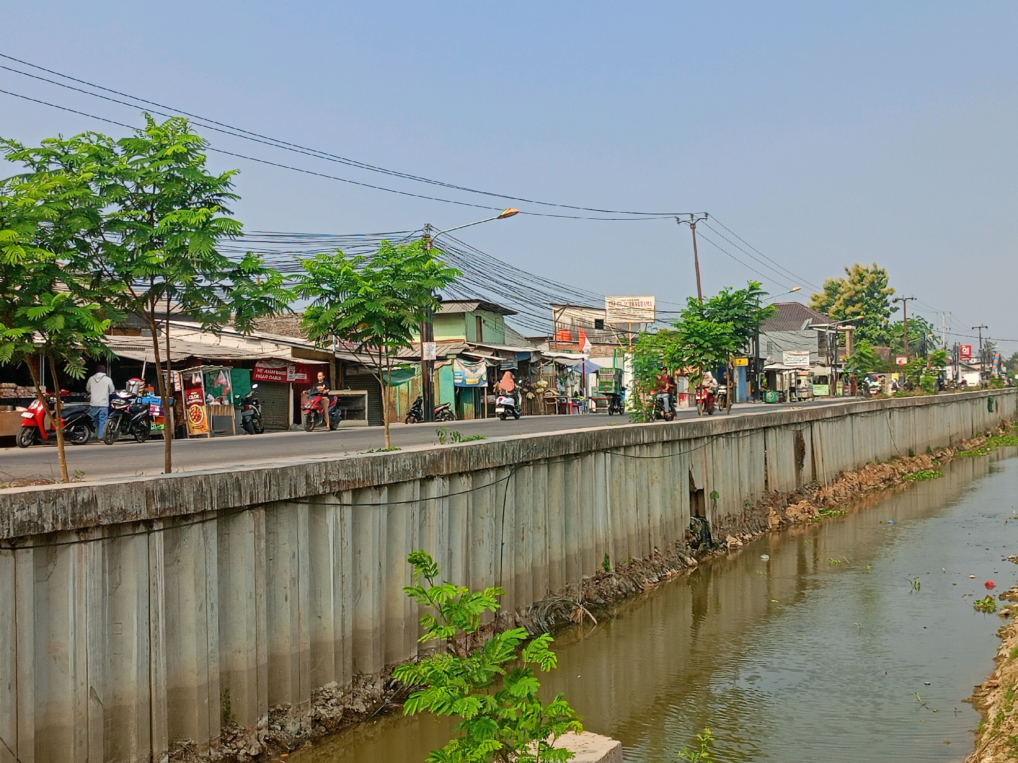 This is a medium-shot, eye-level, exterior photograph of a street scene in Sriamur, Indonesia. The image captures a section of a canal on the right, with a concrete retaining wall separating it from a street on the left. The canal water is murky brown and has some scattered debris and vegetation floating on its surface. The retaining wall is made of vertical concrete pillars and is partially overgrown with small plants at its base.
On the street level, there are several small buildings that appear to be shops or residences, with corrugated metal roofs and various signage. A few trees with green foliage are interspersed along the roadside. Several motorcycles are parked or being ridden on the street. There are a few people visible, mostly on motorcycles or walking near the buildings. The sky is clear and blue, with power lines and a street lamp visible above the buildings. The overall impression is that of a busy, everyday street scene in a developing urban or semi-urban area.