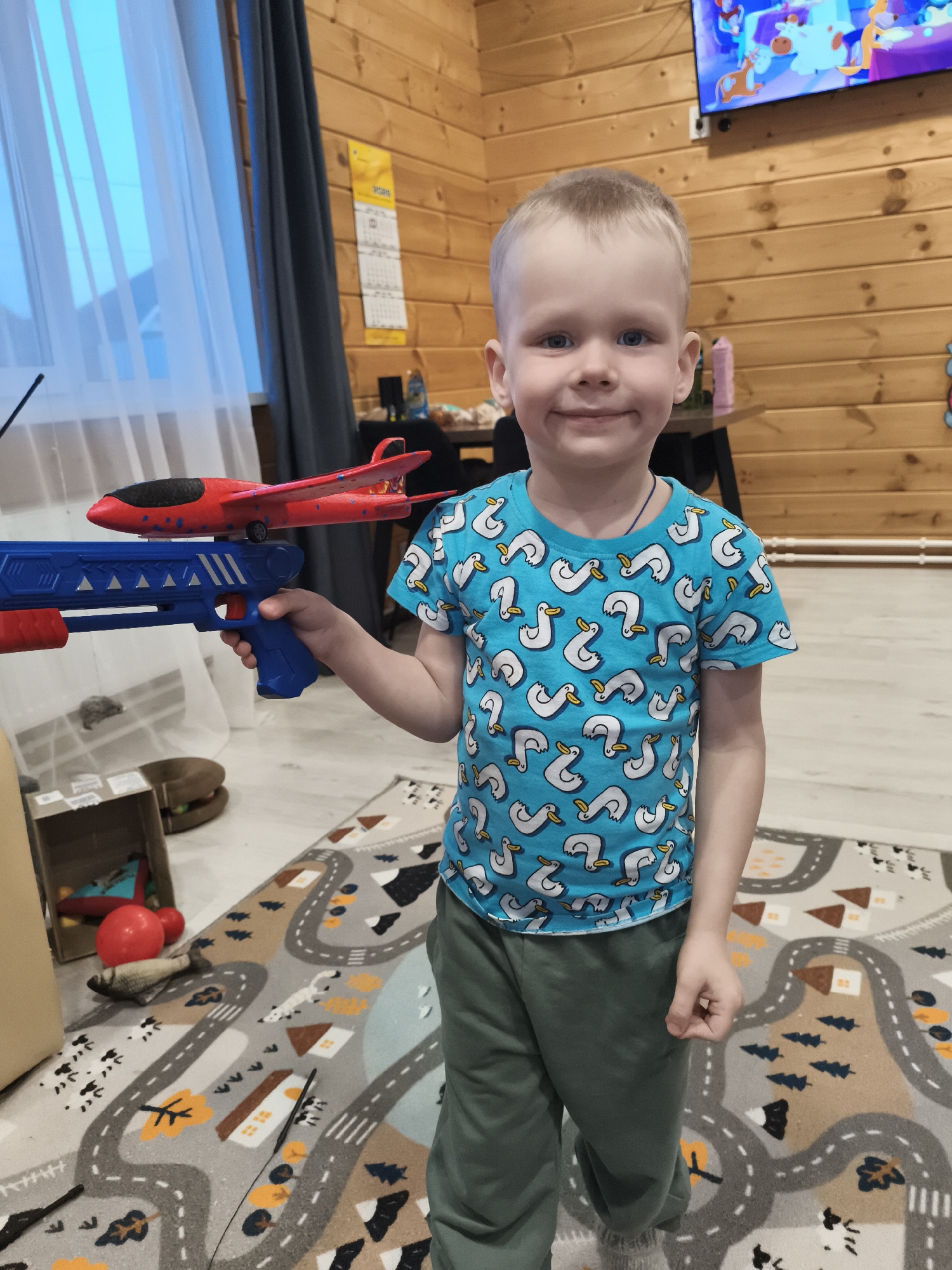 A young boy stands indoors, holding a blue toy gun with a red airplane on top. He is wearing a blue t-shirt with a white duck pattern and green pants. The background includes a carpet with road designs, wooden walls, a window with white curtains, and a television playing a cartoon. The boy is in the city of Русский Вожой, Russia.