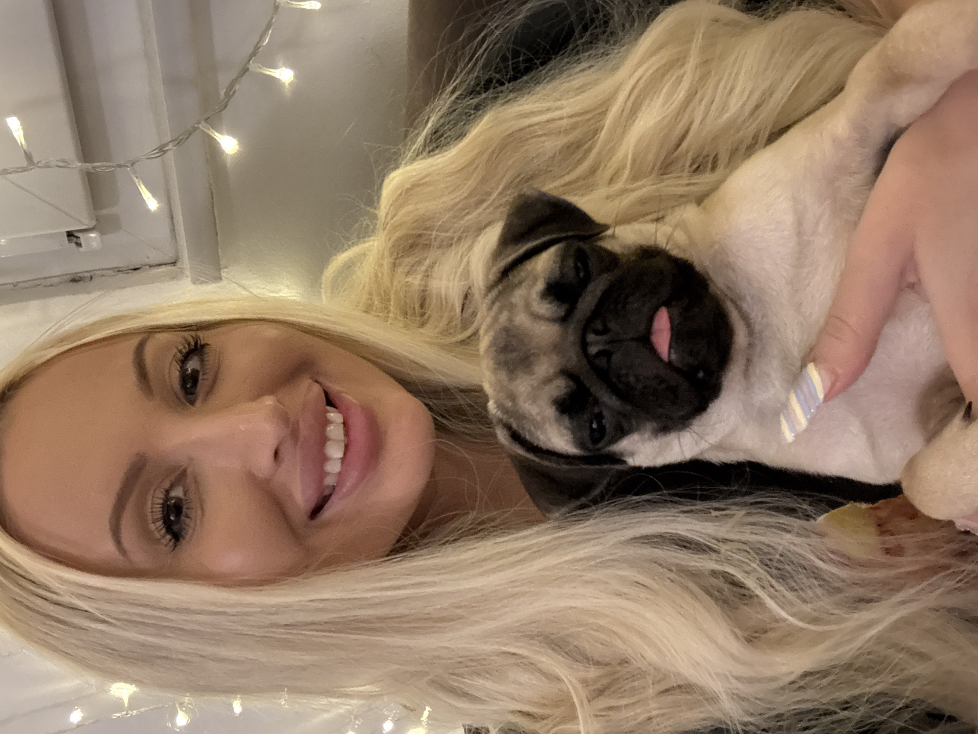 A woman with long blonde hair is holding a pug puppy. The woman is smiling and looking at the camera. The puppy is looking down and has its tongue sticking out. The background is blurred and appears to be indoors, with some string lights visible. The overall lighting is warm and soft. The image appears to be a selfie. The location is Unknown, Unknown.