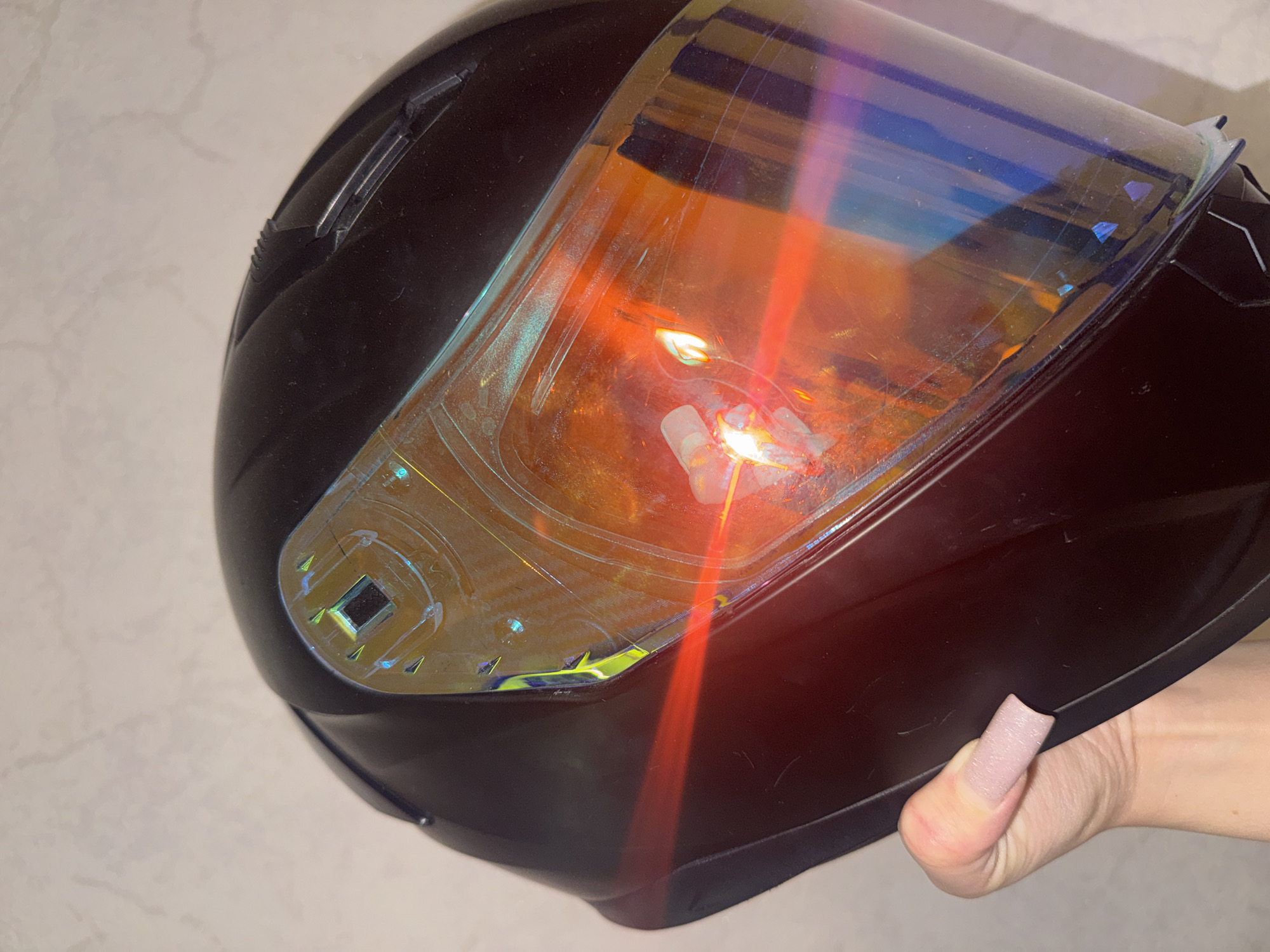 A close-up, slightly angled shot shows a dark helmet with a reflective visor. The visor displays a spectrum of colors, including orange, yellow, blue, and purple, with visible light reflections. The helmet is held by a hand with a light pink, matte fingernail, positioned on the left side of the frame. The background is a neutral, textured wall.