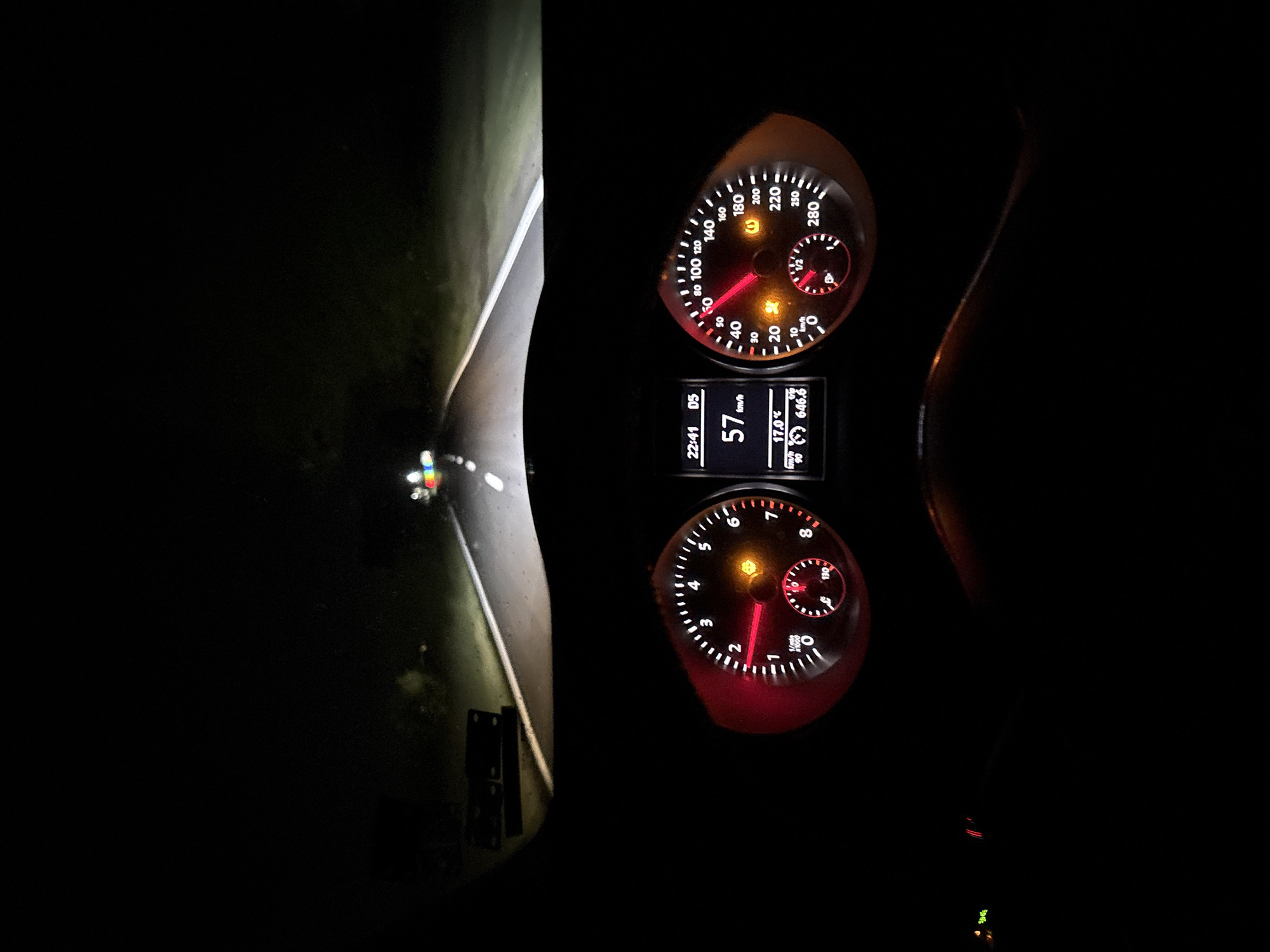 The image captures a night scene from inside a car, showing the illuminated dashboard and the road ahead. The perspective suggests the driver's view, with the dashboard prominent in the foreground and the road stretching into the darkness beyond the windshield.
On the dashboard, two primary analog gauges are visible. The speedometer, on the left, indicates a speed of 57 km/h, with a maximum reading of 280 km/h. A yellow warning light is illuminated within this gauge. To its right, the tachometer shows the engine RPMs around 1500, with a scale extending up to 8 x 1000 RPM. Another yellow warning light is active here.
Below these gauges, a digital display provides additional information: the current time is 22:41 (10:41 PM), the car is in 5th gear ("05"), the speed is reiterated as 57 km/h, the outside temperature is 17.0 °C, and a trip meter reads 646.6 km. A segment of the steering wheel is visible on the right side of the dashboard.
The view through the windshield reveals a dark night road, illuminated by the car's headlights. White lines clearly mark the two lanes of the road, and grassy verges are visible on either side. In the far distance on the left side of the road, faint, multi-colored lights suggest the presence of another object or person, possibly a bicycle with reflective elements or a small vehicle. This scene takes place in Dolna banya, Bulgaria.