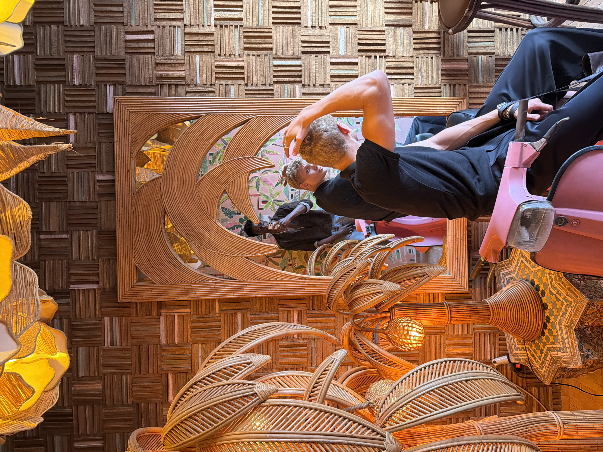 The image depicts an interior scene that appears to be located in Marrakesh, Morocco, likely within a boutique hotel or a themed cafe, given the distinctive decor. The walls are covered in a patterned arrangement of small wooden blocks, creating a textured, warm brown backdrop. Dominating the foreground is a large, ornate mirror with a bamboo-like frame, shaped to resemble palm fronds.
Reflected in the mirror, two young men are visible. One, with blond curly hair and wearing a black t-shirt and black pants, is seen from the back, adjusting his hair with his right hand. He is seated on what appears to be a pink vintage scooter. Another man, with dark skin and wearing a dark shirt, is also reflected, holding a phone up as if taking a selfie.
To the left of the mirror, a large, intricately woven bamboo palm tree sculpture is prominently featured, with its fronds curving upwards and inwards. A rattan palm tree lamp with a woven spherical shade, emitting a warm orange light, is placed on a small, octagonal table covered with a colorful mosaic tile pattern. The table itself rests on the back of the pink scooter. The scooter's front is visible in the lower right of the frame.
The lighting in the scene is warm and ambient, with prominent illumination from the rattan lamp and possibly overhead fixtures, casting a golden glow. The overall aesthetic is tropical and eclectic, with a strong emphasis on natural materials like rattan and wood, and a touch of vibrant color from the scooter and the mosaic table. The setting suggests a stylish and artistic establishment common in tourist-oriented areas of Marrakesh.