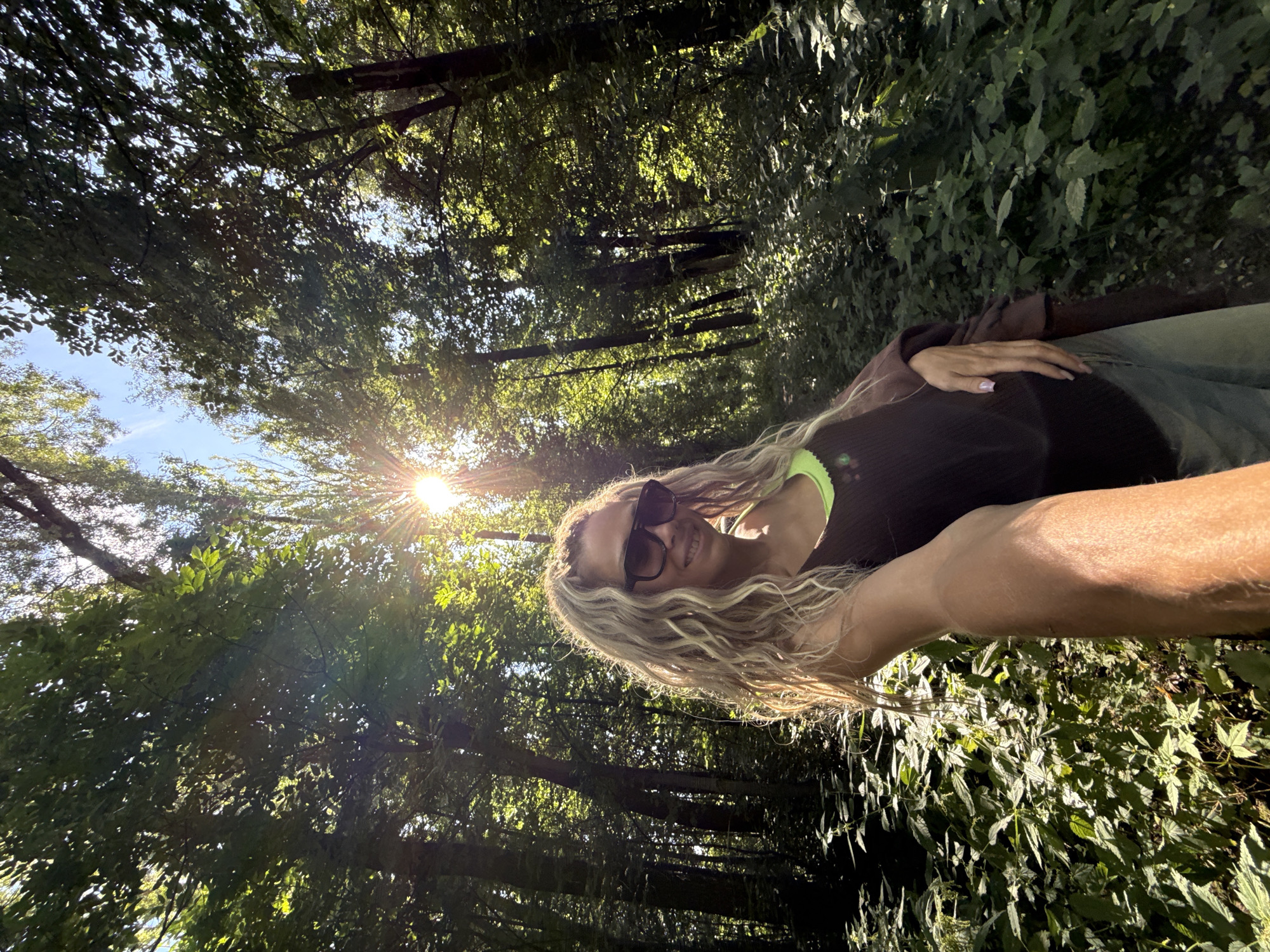 A woman with blonde, curly hair and sunglasses is taking a selfie in a forest. The sun is shining through the trees, creating a dappled light effect. The woman is wearing a dark tank top with a neon green trim and denim jeans. She has a brown jacket draped over her shoulder. The forest floor is covered in lush green foliage. The location is described as Rusanovo, Russia.