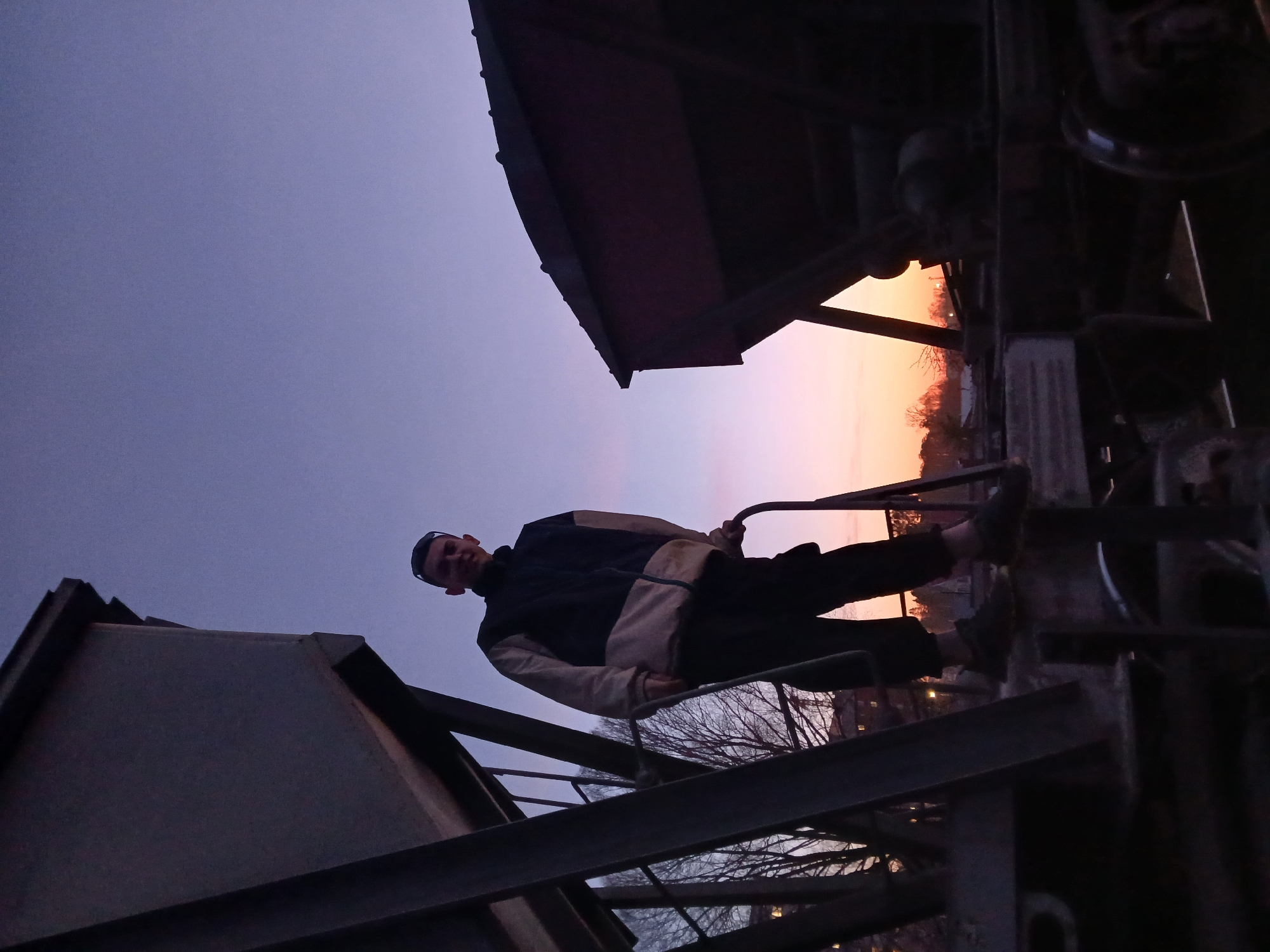The image depicts a young man standing on a large metallic structure during dusk or twilight. The photograph is oriented sideways, with the sky appearing on the left and right, but when viewed upright, the sky forms the background, transitioning from a deep indigo-purple at the top to a warm orange and pink glow near the horizon.
The central subject is a young man, dressed in a dark blue/black and light tan/beige jacket, dark pants, and a dark cap. He is seen holding onto a vertical metal bar, balancing on what appears to be a lower section of the heavy industrial framework, possibly part of a train car or a large piece of machinery. His face is slightly illuminated, and he looks directly at the camera with a subtle, neutral expression.
The setting appears to be an outdoor, possibly industrial or rail yard area, in Chaikovsky, Russia. The metallic structures dominate the foreground and sides, appearing dark and silhouetted against the bright sky. In the mid-ground, the bare branches of trees are visible, suggesting a late autumn or winter scene. The low light conditions accentuate the colorful sky and cast most elements into shadow, creating a dramatic atmosphere. There is no visible text in the image.
