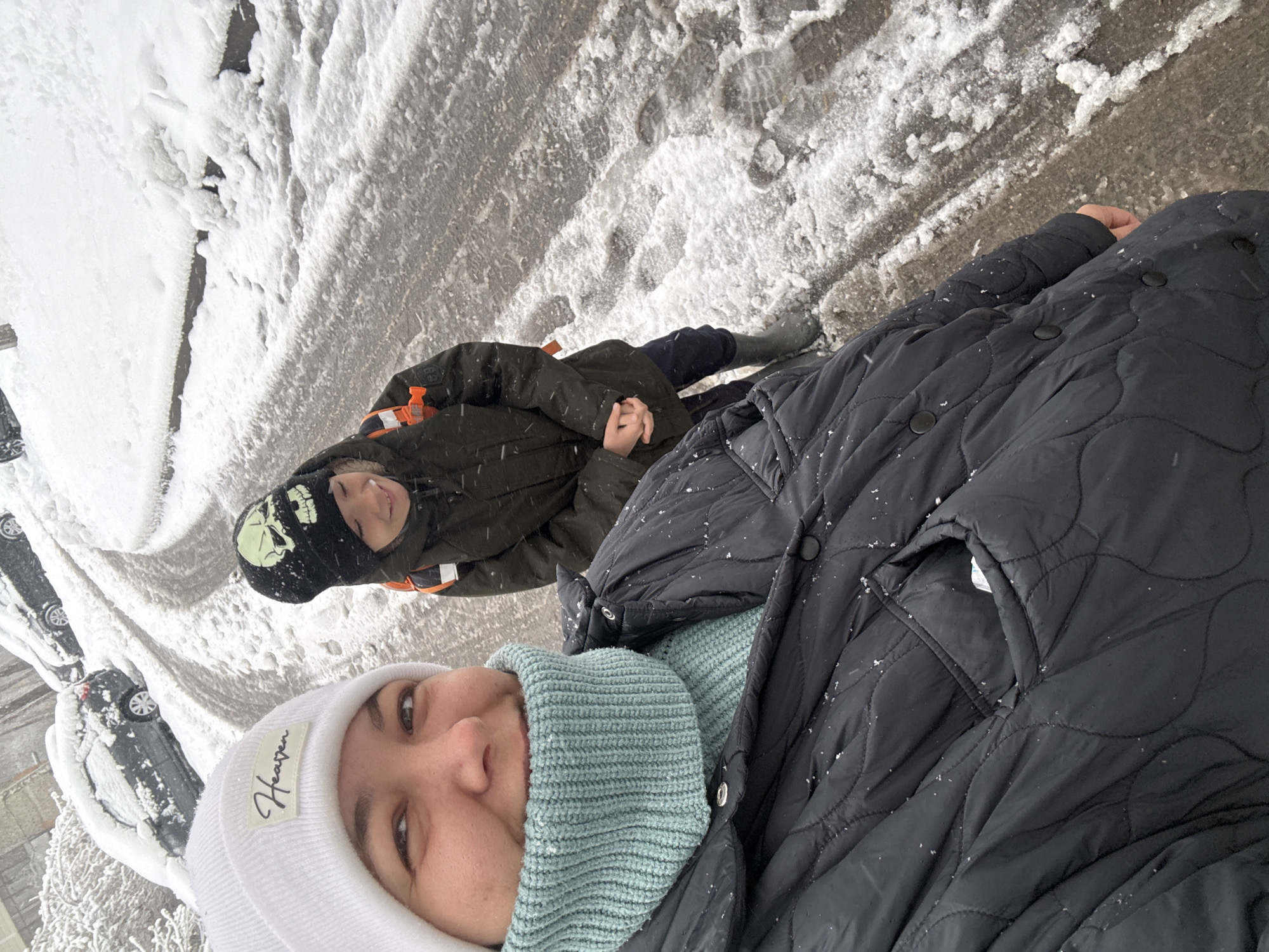 An image captures two individuals, a woman and a child, standing outdoors in a snowy environment in Dmitrov, Russia. The photo is taken from a slightly elevated, selfie-style perspective, with the woman closer to the camera. The ground is covered in fresh snow and slush, showing tire tracks and footprints, suggesting a street or paved area. Several cars, also covered in snow, are visible in the background on what appears to be a residential street.
The woman, in the foreground, is wearing a white knitted beanie with a label, a light blue ribbed neck warmer, and a black quilted winter coat with small snowflakes visible on it. She has a slight smile on her face. The child, further back, wears a dark green winter coat with an orange attachment, dark pants, and a black beanie adorned with a glow-in-the-dark skull design. The child looks up towards the camera with a neutral expression.
The weather is clearly snowy and cold, indicated by the snow on the ground, cars, and the individuals' winter attire, as well as the visible snowflakes. The lighting suggests daytime under an overcast sky.
A notable detail is the white fabric label on the woman's beanie, which reads "Heaven". The overall scene depicts a winter day, possibly during a snowfall, with the two individuals bundled up for the cold.
