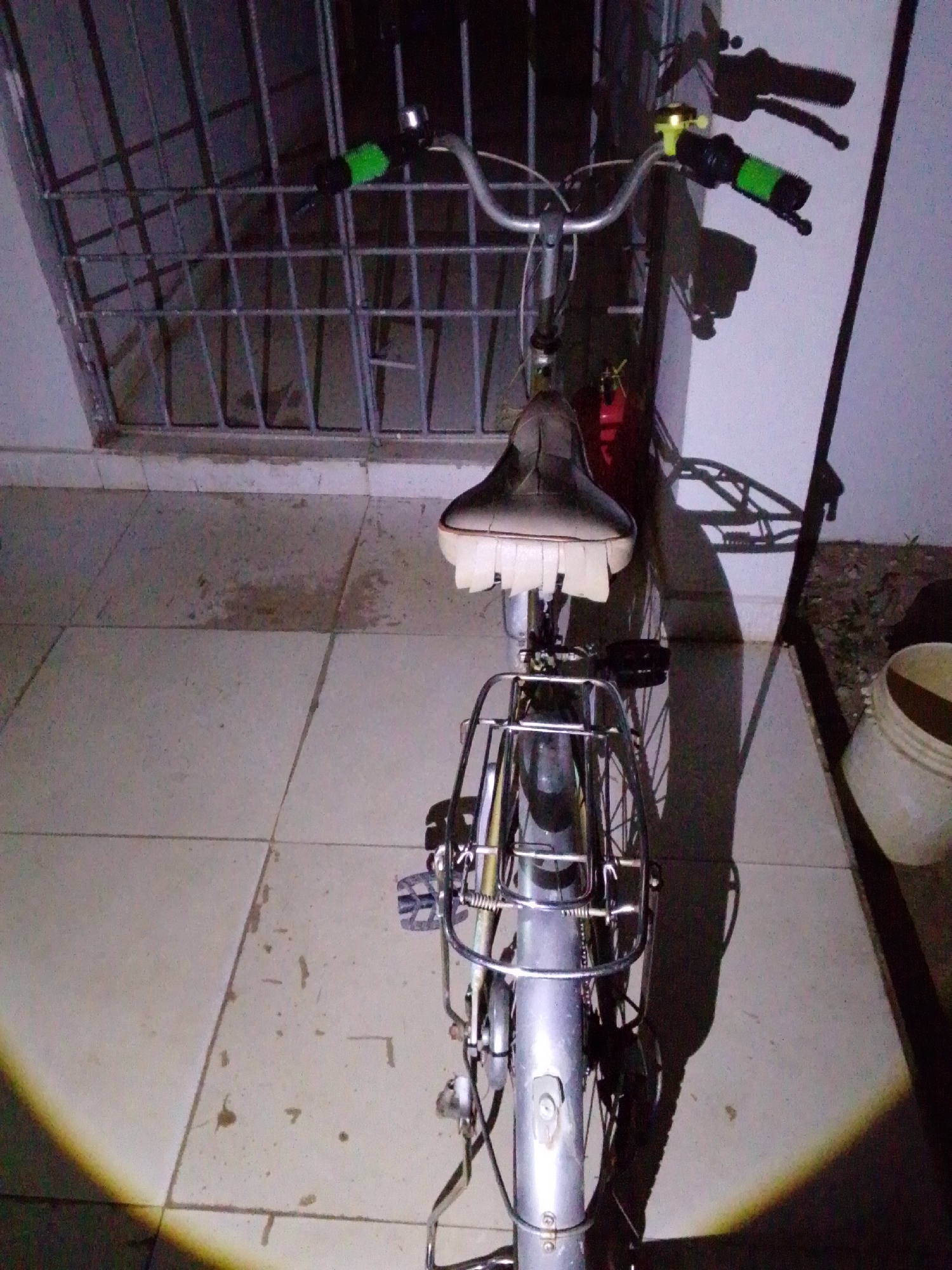 A low-angle, full-shot photograph taken at night captures a silver bicycle parked on a tiled floor in Serti, Nigeria. The bicycle, positioned facing forward, has a black seat and a silver frame with a metal basket on the rear. Green handlebar grips and a yellow bell are visible on the handlebars. Behind the bicycle, a metal grate covers a window or opening, and to the right, a white bucket sits on the ground. The scene is illuminated by artificial light, casting shadows on the wall and floor. The ground is composed of light-colored square tiles, with some dark stains or marks present.