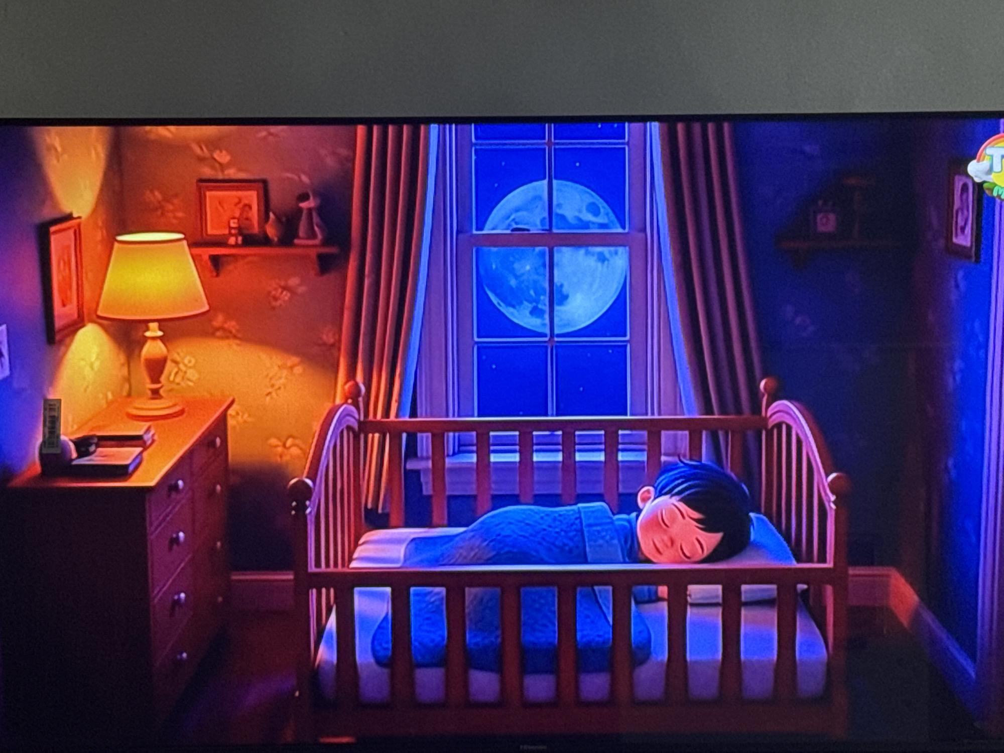 The image displays a bedroom scene with a child sleeping peacefully in a wooden crib. The room is dimly lit, with a large, full moon visible through a window. The moon's light casts a blue glow on the room, contrasting with the warm, orange light emanating from a bedside lamp on a wooden dresser. The dresser has several drawers with silver knobs and is adorned with two framed pictures and a small decorative figure on the top. The walls are covered in a patterned wallpaper, and red curtains frame the window. The child is covered with a blue blanket and appears to be dreaming. The scene evokes a sense of calm and tranquility, suggesting it is nighttime. There is no discernible connection to the city of Chardara, Uzbekistan.
