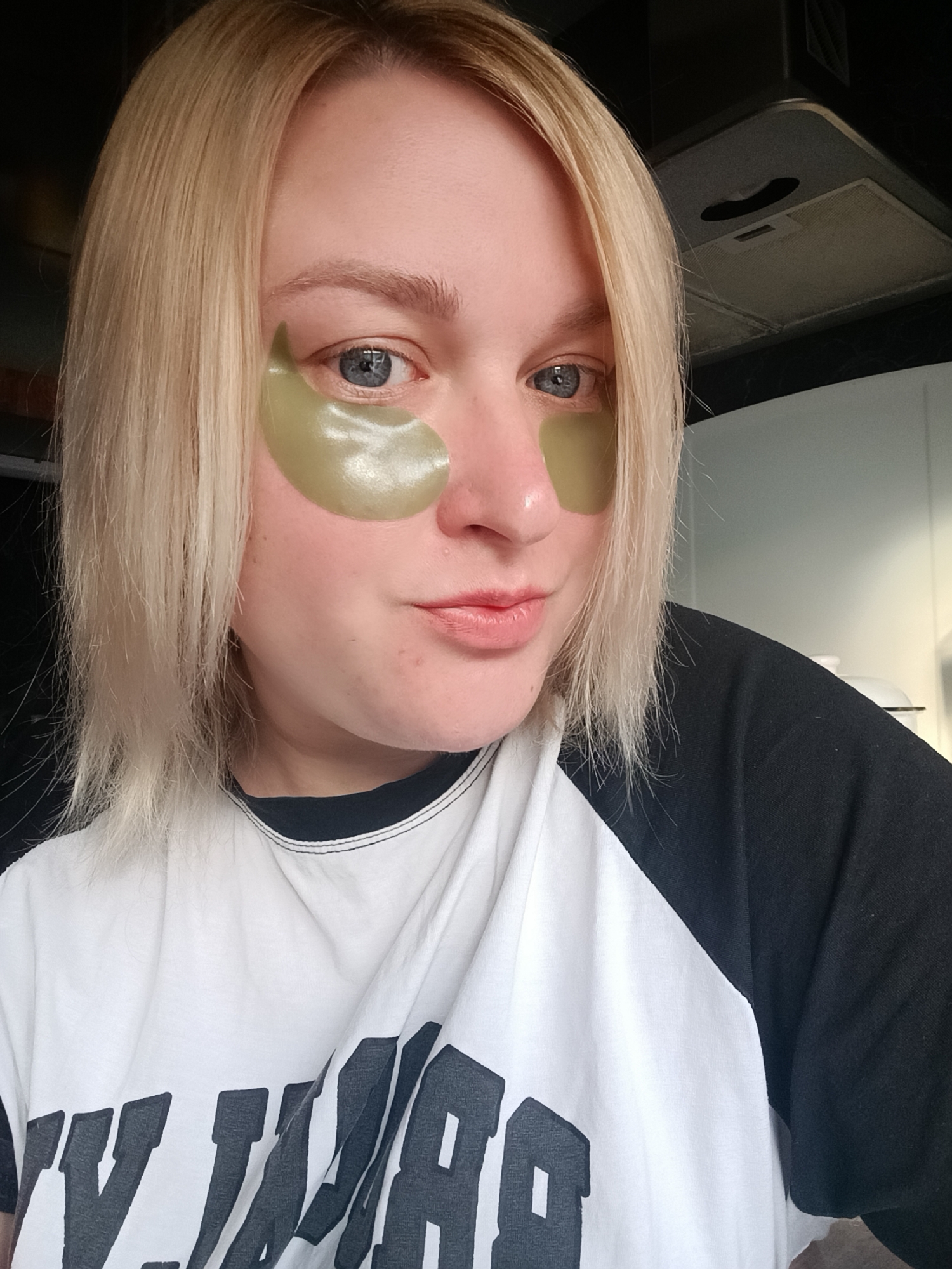 A woman with light blonde shoulder-length hair and blue eyes is the central figure in this selfie. She is wearing green eye patches under each eye. The woman is wearing a white t-shirt with black raglan sleeves and a black graphic print across the front. The background is partially visible, showing what appears to be a kitchen setting with a range hood above a stove and a partially visible white appliance. The location is Chaikovsky, Russia.