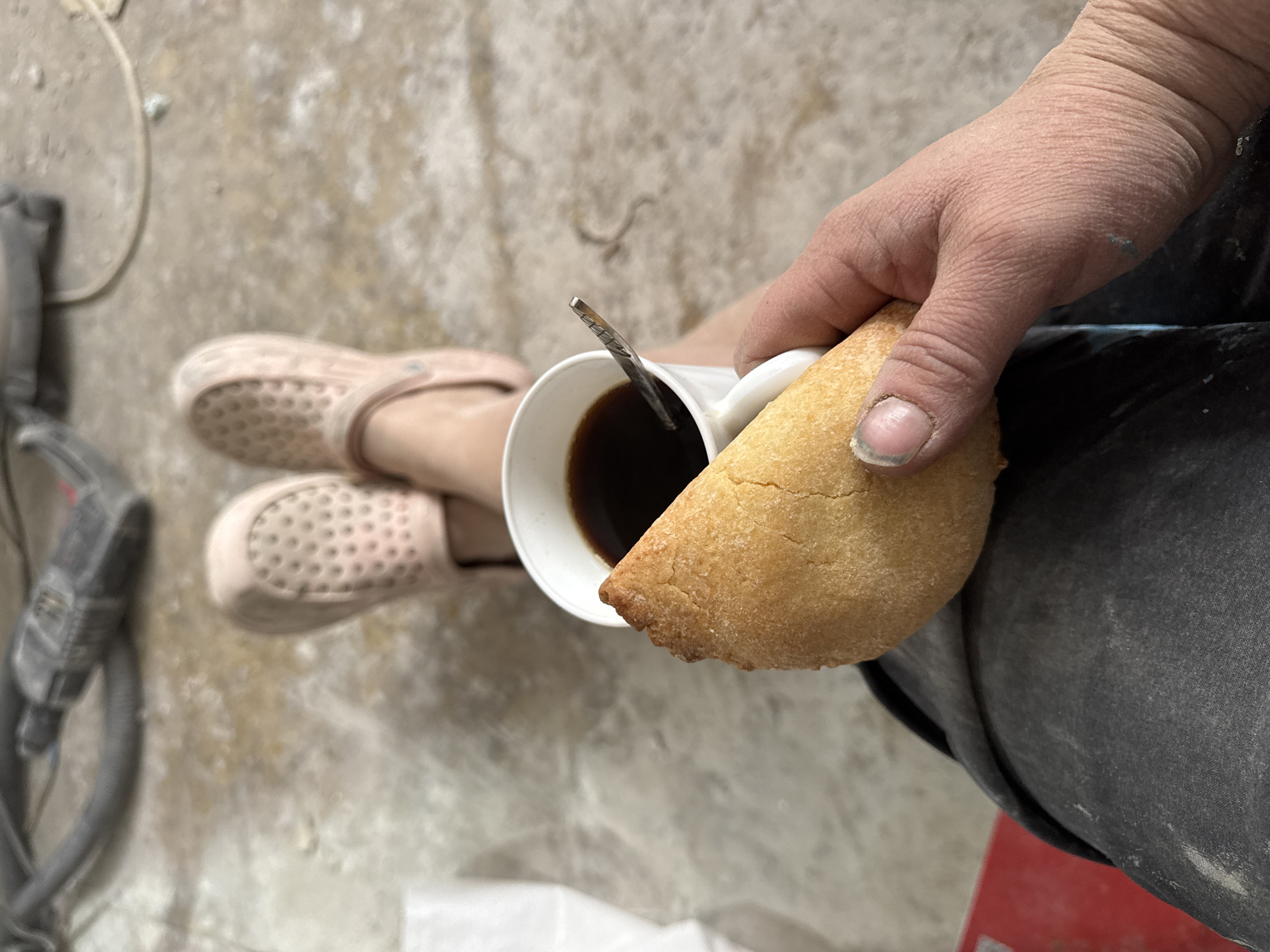 The image shows a person sitting down, holding a cup of black coffee and a triangular pastry. The person is wearing pink clogs. To the left of the feet is a vacuum cleaner. The pants the person is wearing appear to be grey and dirty. The background is a light grey concrete floor. There is no information in the provided image to confirm the location as Chaikovsky, Russia.