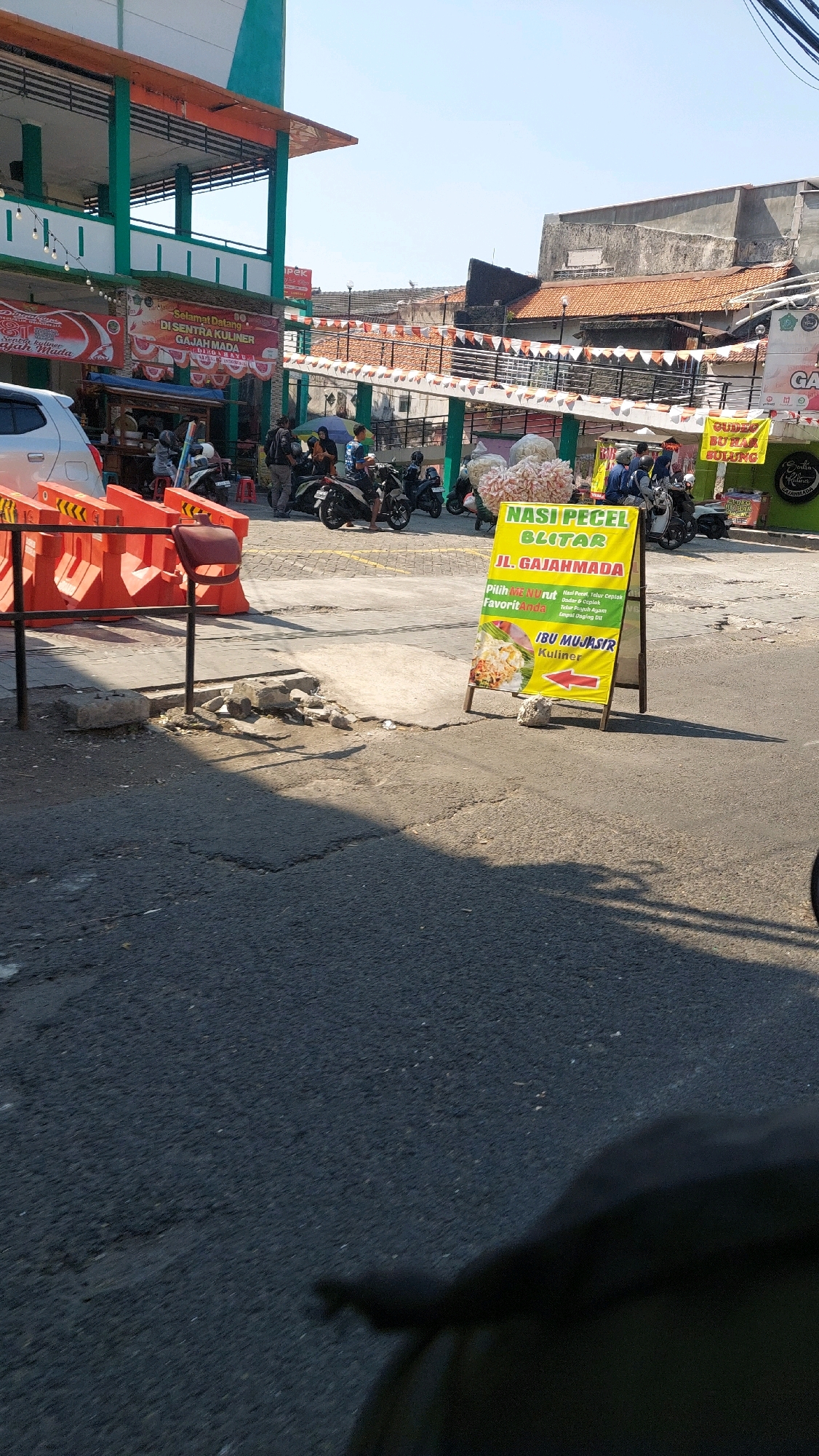 The image depicts a street scene in Sidoarjo, Indonesia, likely at a food stall or market. A yellow A-frame sign is prominently displayed, advertising "Nasi Pecel Blitar Jl. Gajahmada". The sign also lists menu options such as "Nasi Pecel", "Telur Ceplok", "Dadar / Omelet", "Tahu / Tempe", "Rujak Ayam", and "Empal Daging". Below these options, it says "IBU MUJIASTRI Kuliner".
In the background, a building with green and orange accents is visible, possibly a market or food court. Several motorcycles are parked, and a few people are gathered around. Orange traffic barriers are placed on the side of the road. The environment appears to be outdoors, with a clear sky and sunlight casting shadows on the asphalt. A white car is partially visible on the left. The ground is paved with asphalt, showing some cracks and patches. In the foreground, a dark object, possibly a person or vehicle in shadow, occupies the bottom right portion of the image. The overall impression is of a busy, everyday scene in a commercial area.