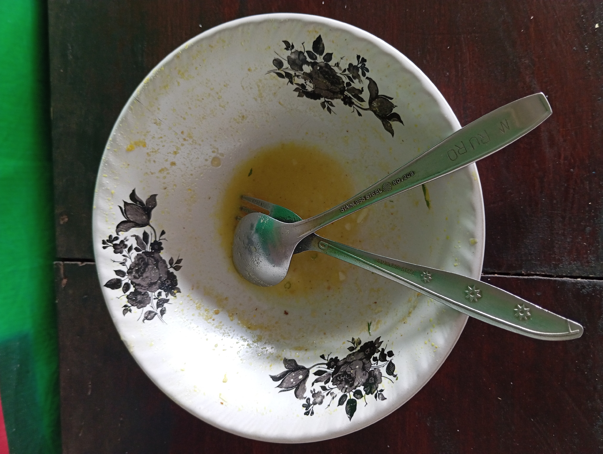An overhead shot shows a dirty white plate with black floral designs on a dark wooden table. The plate contains remnants of food, appearing as yellow residue. A fork and a spoon are crossed in the center of the plate, with their handles extending out. The fork's handle is inscribed with "STAINLESS STEEL NO. 203" and the spoon's handle is inscribed with "M RURO". The background shows a green surface above the table. The scene is indoors, likely a domestic setting in Lemahbang, Indonesia.