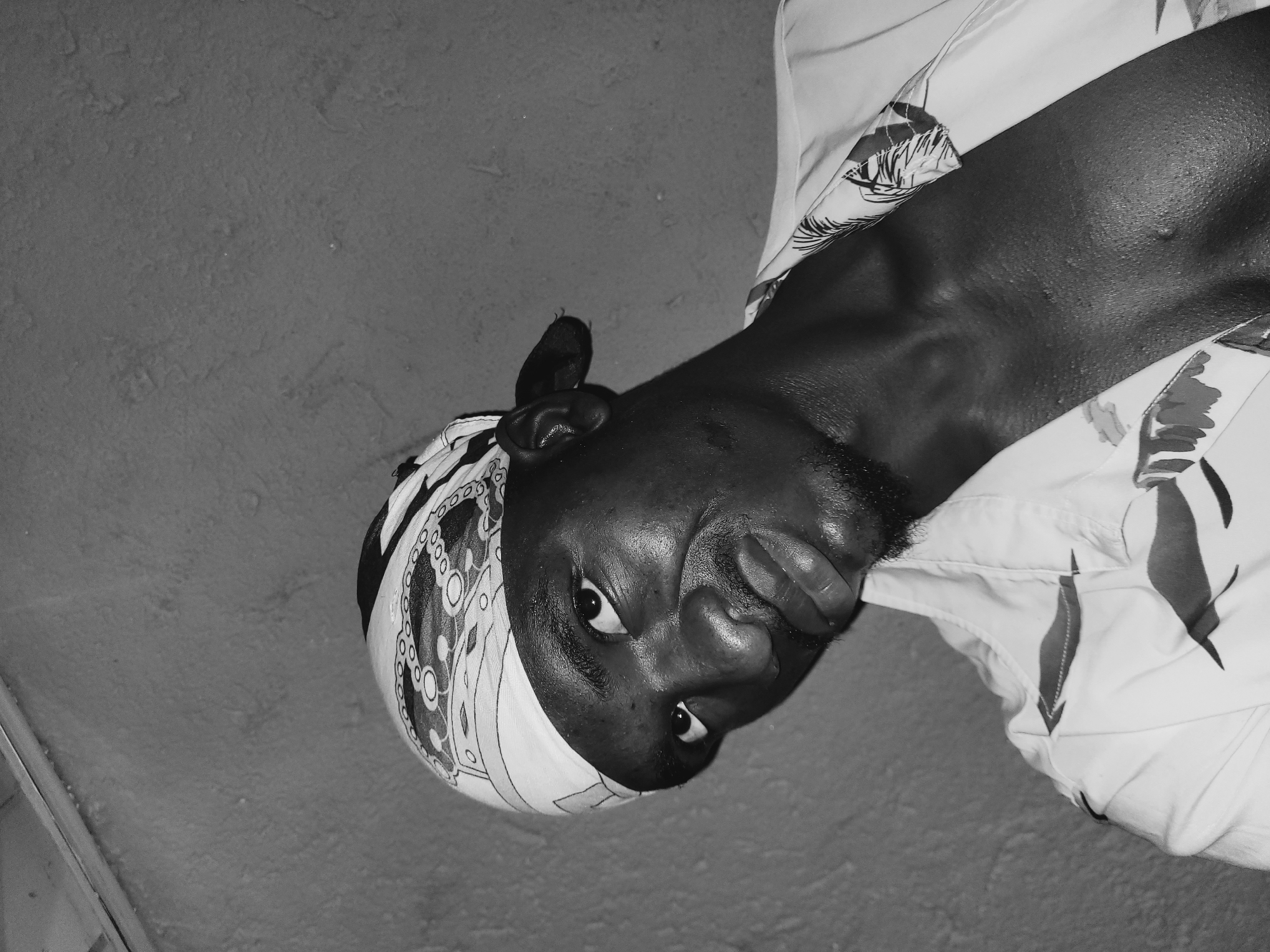 A black and white, waist-up portrait shows a Black man wearing a patterned bandana and a tropical-print button-down shirt, open at the neck. He is looking up and to his right, with his head tilted to the left. His skin is glistening, suggesting sweat. The background is a textured, neutral-colored wall. The image appears to be taken indoors.