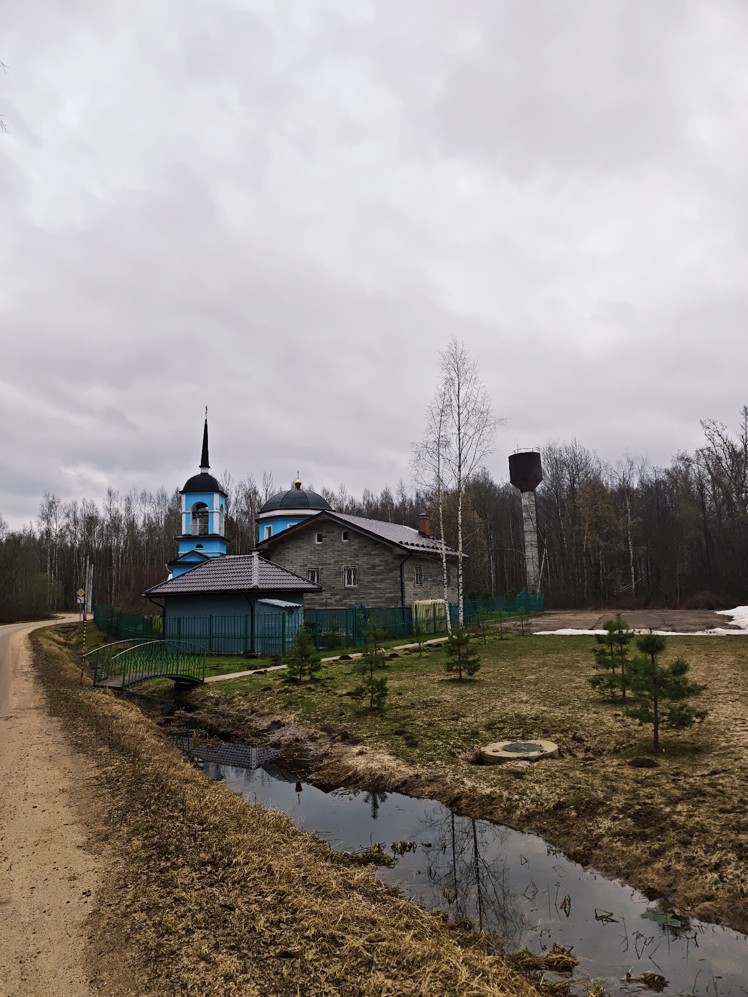 The image captures a landscape in Nadezhdino, Russia, under a cloudy sky. A road runs along the left side of the frame. A stream flows parallel to the road, reflecting the surrounding structures and trees. A small green bridge crosses the stream.
A church complex is visible beyond the stream, enclosed by a green fence. The complex includes a blue and black bell tower and a gray building with a brown roof. A water tower is located further back, partially obscured by trees. The ground is covered with brown grass, with patches of snow in the distance. Young pine trees are scattered across the area.