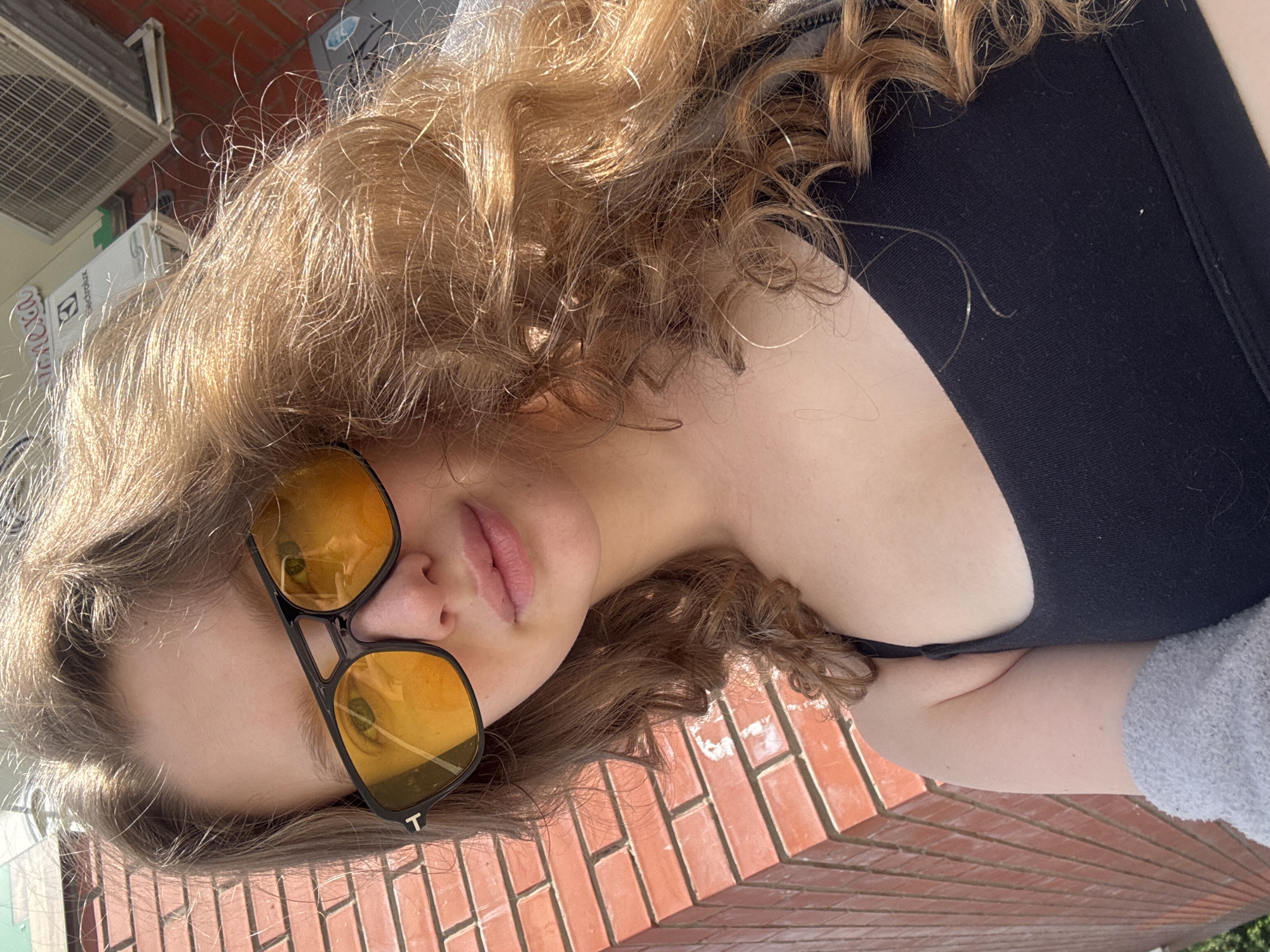 The image shows a close-up of a young woman with long, curly, light brown hair, wearing large, dark-framed sunglasses with orange lenses. She is wearing a black sports bra and a light gray fuzzy sleeve on her left arm. The background consists of a red brick wall to the left and a building with an air conditioning unit and signs to the right, suggesting an outdoor urban setting. Based on the visual clues, the location appears to be in the city of Izhevsk, Russia.