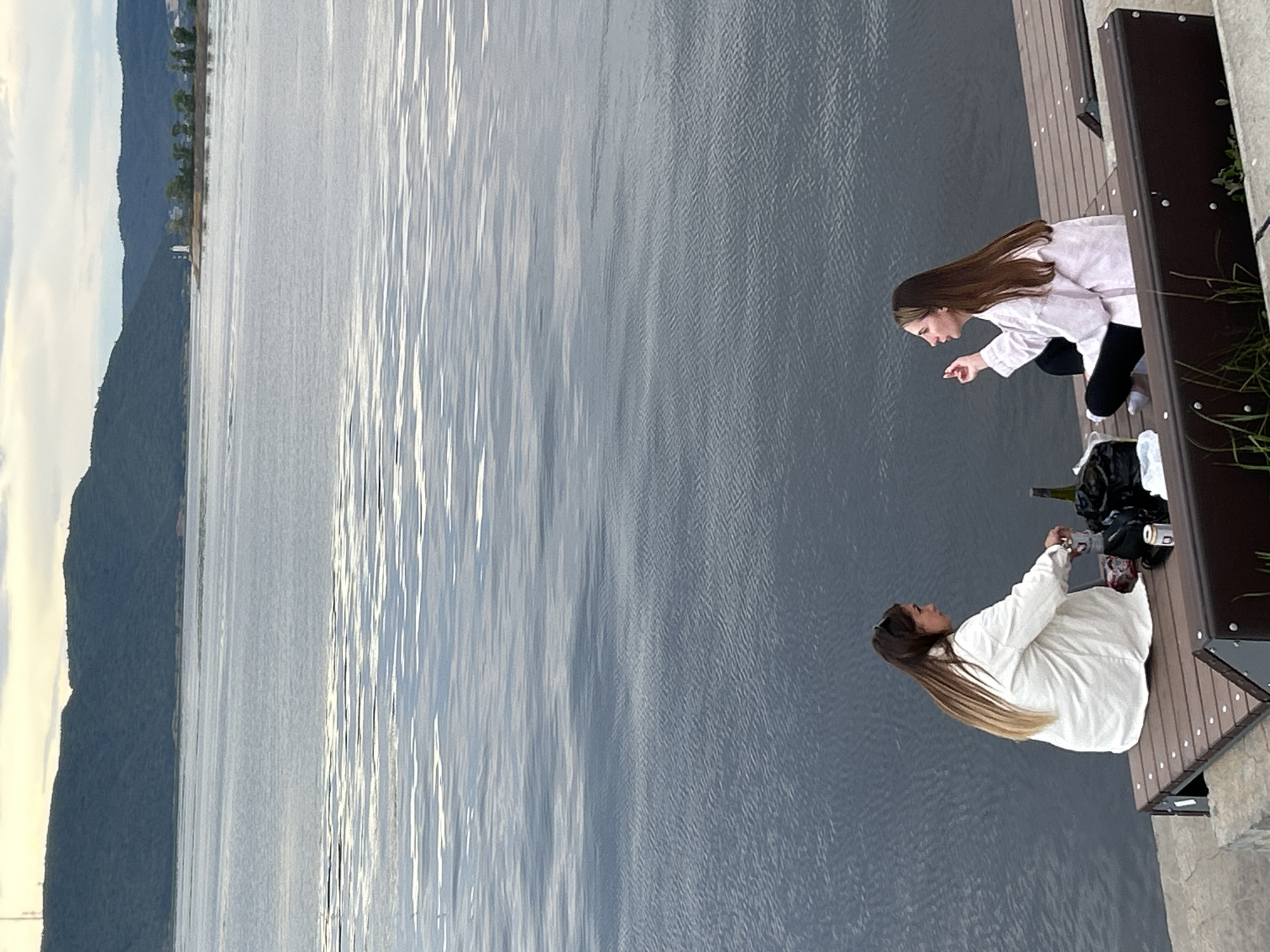 Two young women are having a picnic on a wooden deck overlooking a body of water, possibly the Volga River, near Tolyatti, Russia. The woman on the right is wearing a light pink hoodie and black pants, and is crouched down, pointing at something in the water. The woman on the left is wearing a white jacket and is also crouched down, looking in the same direction. There are drinks and snacks on the deck, including a bottle of wine and some cans. The sun is setting, casting a warm glow on the water and the surrounding landscape, which includes a forested hill. The mood appears relaxed and enjoyable. The time of day is late afternoon or early evening. The weather appears to be clear and calm.