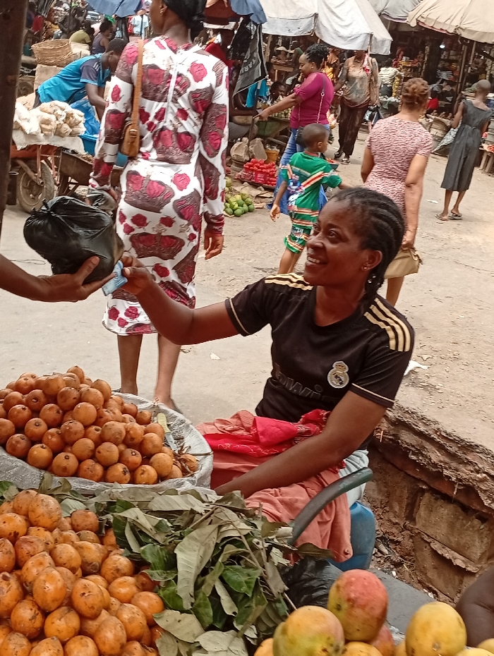 This image captures a vibrant and busy outdoor market scene, likely in a tropical or subtropical region, centered around a vendor interacting with a customer.
In the foreground, to the right, a woman with dark braided hair is seated, looking towards the left with a warm smile. She is wearing a black short-sleeved t-shirt with gold stripes on the sleeves and a white logo on the chest, which appears to be the Real Madrid football club crest. Her right hand is extended, holding paper currency, seemingly in the process of a transaction. On the left, partially visible, another person's hand is holding a black plastic bag and more paper currency, meeting the vendor's hand.
Spread across the bottom of the frame are large quantities of fresh fruit, likely the vendor's merchandise. Two large basins or containers hold piles of small, round, yellowish-orange fruits, which resemble guavas. Mixed among these fruits are fresh green leaves. In the very bottom right corner, a few larger, reddish-yellow fruits, identifiable as mangoes, are also visible.
The background reveals a bustling market environment. Numerous people are walking around, suggesting a busy day. In the mid-ground, a woman wearing a distinctive white dress with red or maroon crown patterns is seen from behind, walking away from the camera. Further into the background, a young child in a green and white striped outfit is visible. Overhead, various tarpaulins or awnings provide shade for the market stalls and pathways. Carts, baskets, and other goods are scattered throughout the background, indicating a diverse array of items being sold. The ground appears to be unpaved, possibly dirt or gravel.
The overall atmosphere suggests an active commercial hub, with the smiling vendor indicating a positive interaction within the busy market setting. The daylight suggests it's daytime.