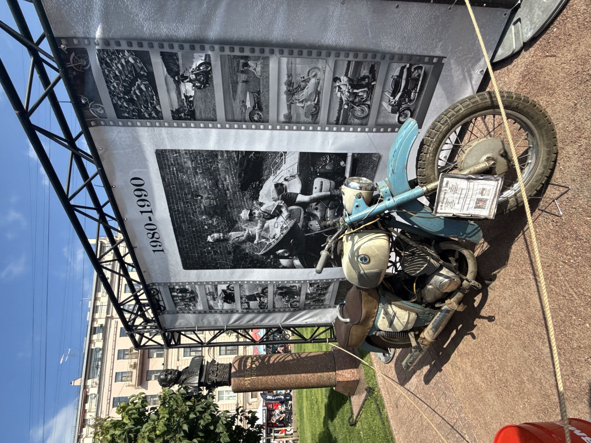 The image shows a vintage motorcycle displayed outdoors, likely at a public event or exhibition. The motorcycle is a light blue and cream color, with a brown seat and saddlebags. It is positioned on a red brick paved area, with a grassy area and a building visible in the background. To the left of the motorcycle, there is a bust on a tall, granite pedestal, with some text inscribed on it. Behind the motorcycle, there is a large banner displaying black and white photographs of motorcycles and people from the years 1980-1990. The overall setting suggests an outdoor exhibition or historical display in a city. The architecture of the building in the background, with its arched windows and balconies, is characteristic of Saint Petersburg. The presence of what appears to be a monument or statue, and a large banner for an exhibition, further supports this.