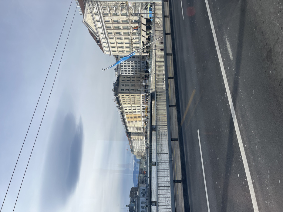 This media file captures a wide urban waterside landscape in Geneva, Switzerland, viewed from a slightly elevated perspective, possibly through a window with significant reflections. The image is taken at daytime under a bright, mostly clear sky with some elongated, wispy clouds.
In the foreground, an asphalt road with white and yellow lane markings is visible, separated by a light-colored barrier or railing from what appears to be a pedestrian walkway or another structure. This suggests the photo might be taken from a bridge or a vehicle on one.
Beyond the railing, a large body of water (Lake Geneva) stretches out, reflecting the sky and distant elements. On the far side of the water, a long row of multi-story buildings with classical architecture lines the waterfront. Many of these buildings feature numerous windows, balconies, and some have awnings or scaffolding. A light blue flag flies from a pole in front of one of the buildings. Distant mountains, possibly the Alps, are visible on the horizon, partially obscured by reflections on the viewing pane.
Notable details include two dark overhead wires cutting across the top left portion of the sky. The most prominent reflection on the viewing surface is a dark, blurred oval shape in the sky area, and a faint reflection of buildings and mountains in the lower half of the image, superimposed over the actual water and land. Text on the scaffolding of one of the buildings is barely legible, possibly indicating "LES BERGUES" or parts of a hotel or cafe name. No explicit people are discernible in the scene.