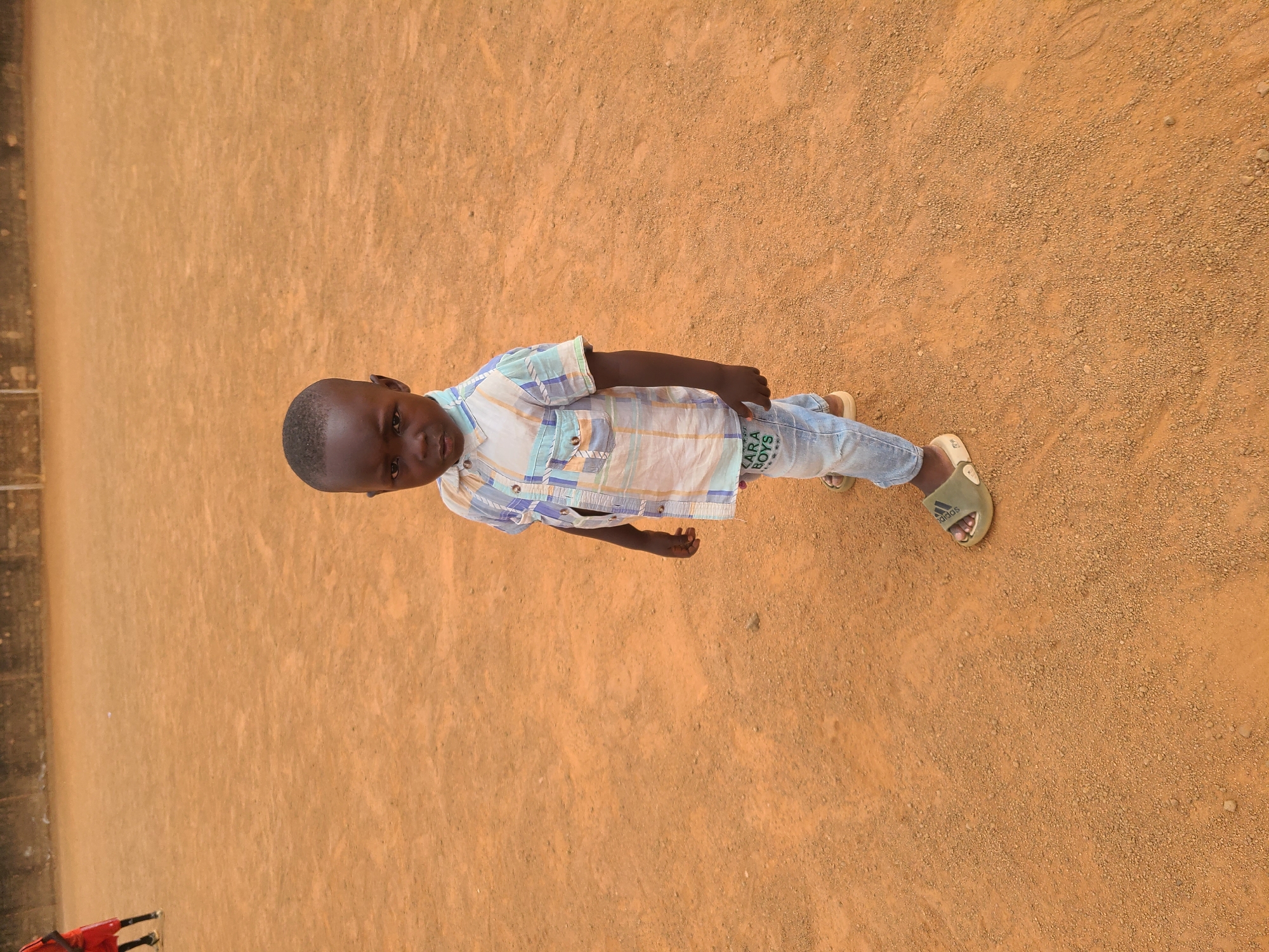 A young boy stands on a reddish-brown dirt surface in Barakin Lerawi, Nigeria. He wears light-blue jeans, a white T-shirt with a design, and a plaid button-down shirt. He also has Adidas-branded footwear.