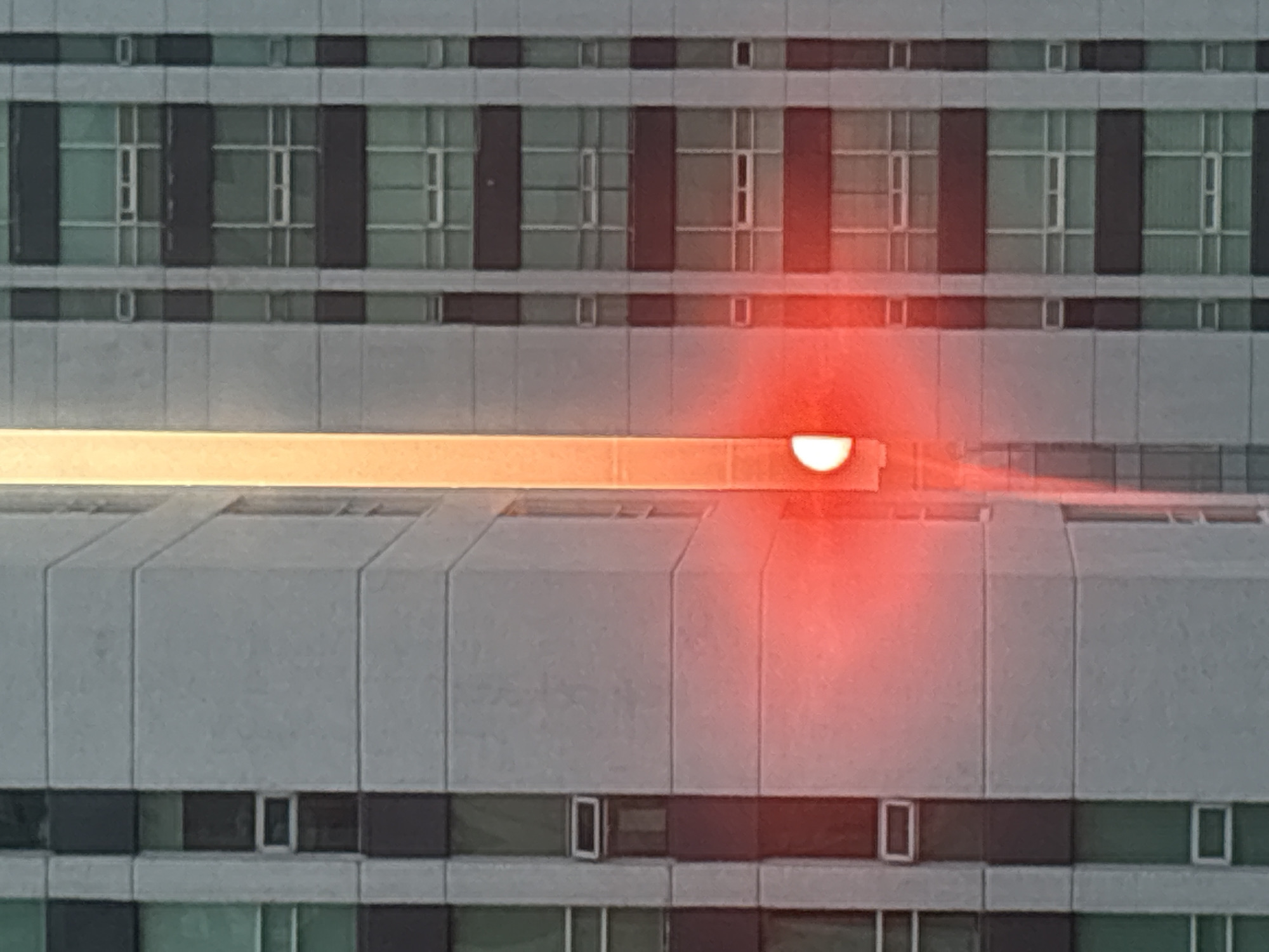 A mid-level, head-on shot of a multi-story building, potentially a hospital or office complex, in daylight. The building's facade features a repetitive pattern of windows, separated by grey panels. A horizontal, orange glow is centered on the image, emanating from behind one of the building's structures, which casts a slight shadow downwards. An area of red light is cast around and below the orange light. The location is potentially Jesús del Monte, Mexico.