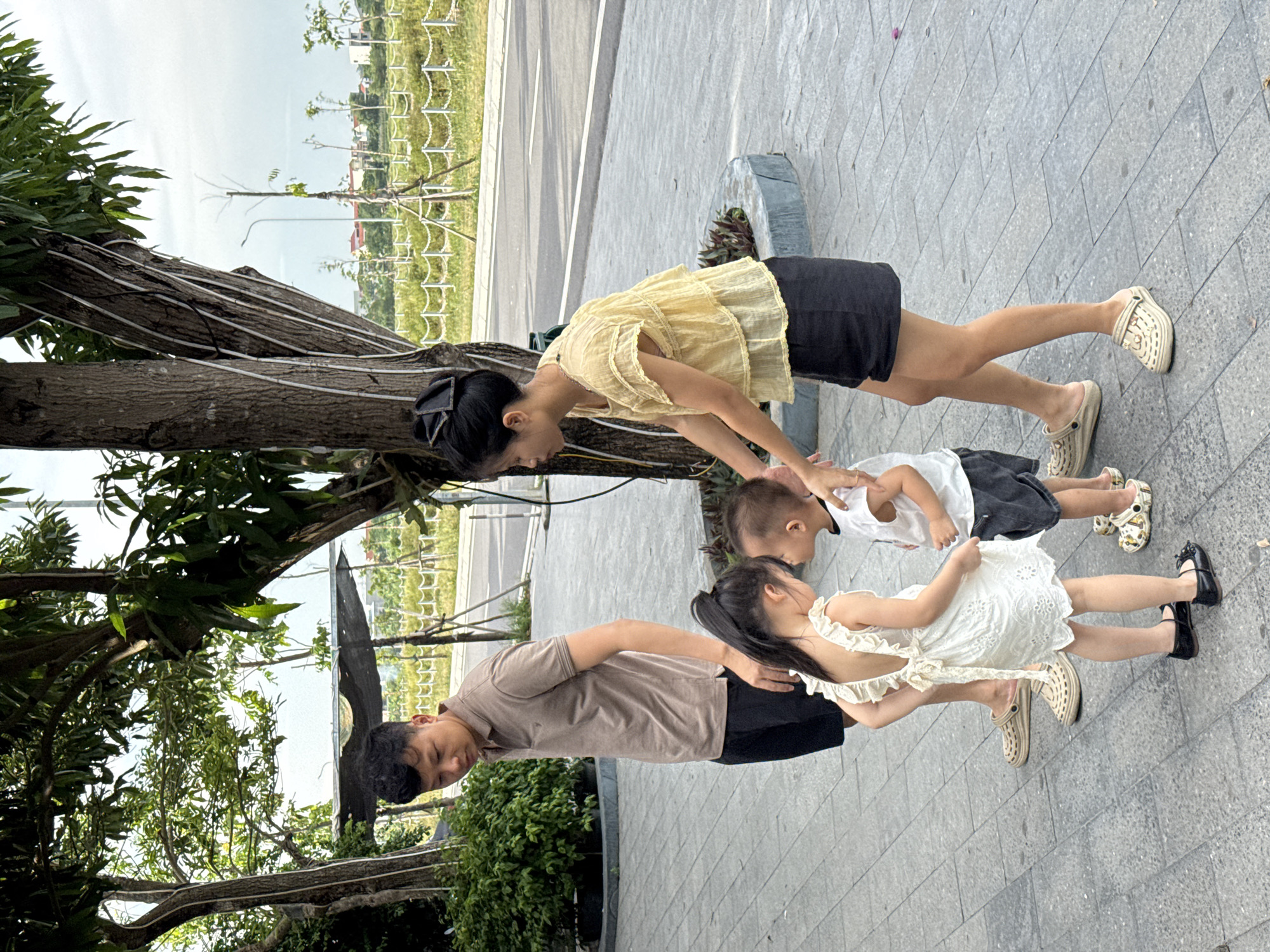 In Hoa Lư Ward, Vietnam, four individuals, two adults and two children, are positioned on a paved area near a street. A man in a tan polo shirt and black shorts stands to the left, looking down. A woman in a yellow ruffled blouse and black shorts is to his right, interacting with the children. The two children, a girl in a white dress and a boy in a white t-shirt and dark shorts, are facing each other. The girl has her arm around the boy. They are all barefoot or wearing casual shoes. The background includes trees, a road, and distant buildings under an overcast sky.