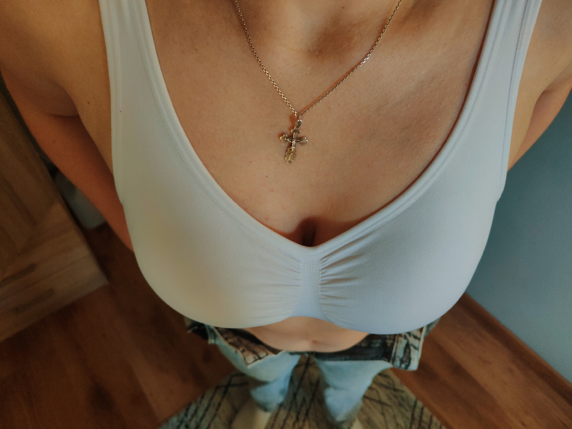 This is a medium shot, eye-level, interior photograph of a person wearing a white bra and blue jeans. The person is wearing a silver cross necklace. The background consists of a wooden floor and a blue wall. The photograph does not contain any information that would allow for its location to be determined as Nowy Dwór Mazowiecki, Poland.