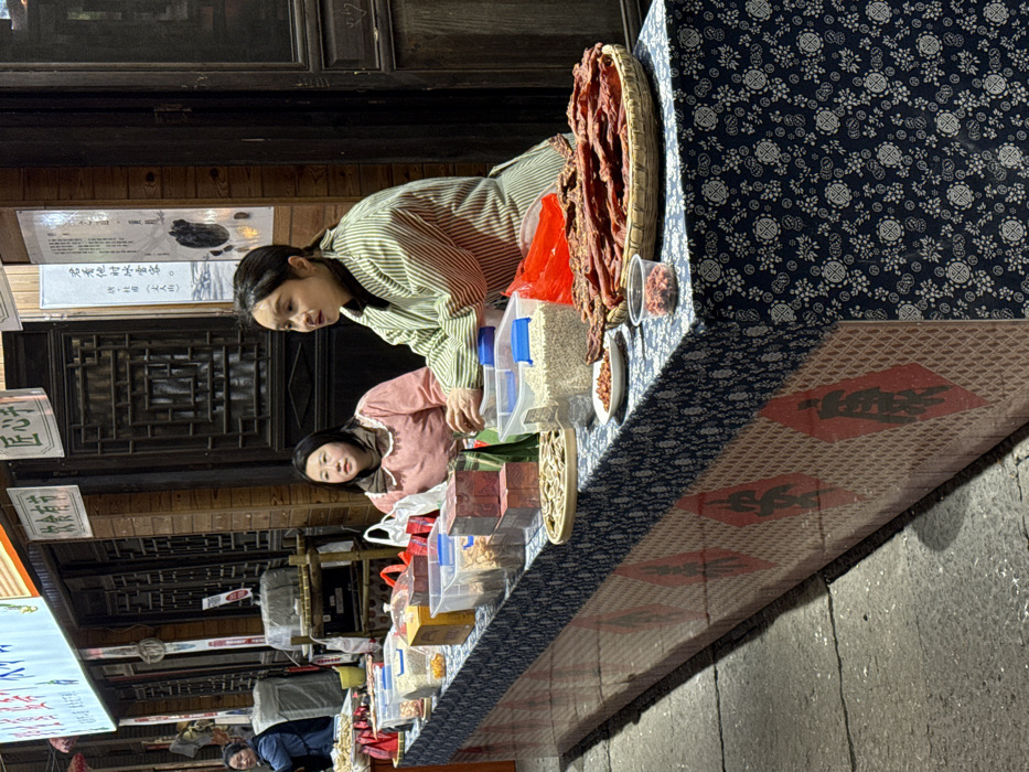 This image depicts a street market stall in Pengshui Miao and Tujia Autonomous County, China, likely during evening hours. Two women are seated behind a long, rectangular display table laden with various food products. The table is covered with a dark blue and white floral-patterned tablecloth, with the lower front section featuring a red checkered design interspersed with large red diamond shapes containing black Chinese characters.
The woman on the right has dark hair pulled back and wears a light green and white striped long-sleeved top, possibly an apron, over a darker garment. Her hands rest on the table near a clear plastic container. The woman on the left, with dark hair, wears a light pink long-sleeved top with a white frilled collar. Both women are looking forward, appearing to attend to the stall. A third woman in a blue jacket is partially visible further down the street to the far left.
The stall displays numerous food items, including various dried goods, nuts, and seeds contained in clear plastic boxes and containers. Several red wooden boxes are also present. On the right, a large quantity of reddish-brown dried meat strips (possibly jerky) is arranged in a flat, round woven basket. Small white dishes and a clear plastic cup hold samples of nuts or dried berries. A flat, round woven tray near the center holds light-colored dried food items, possibly mushrooms or vegetables.
The background consists of dark wooden storefronts with intricate lattice-style windows and doors. Several signs with Chinese characters are visible above and on the buildings, including "饮食有节" and "匠心手." A vertical white banner with Chinese script and an illustration hangs on the wall behind the right woman. The foreground shows grey paving stones on the ground. The scene is illuminated by artificial overhead lighting.