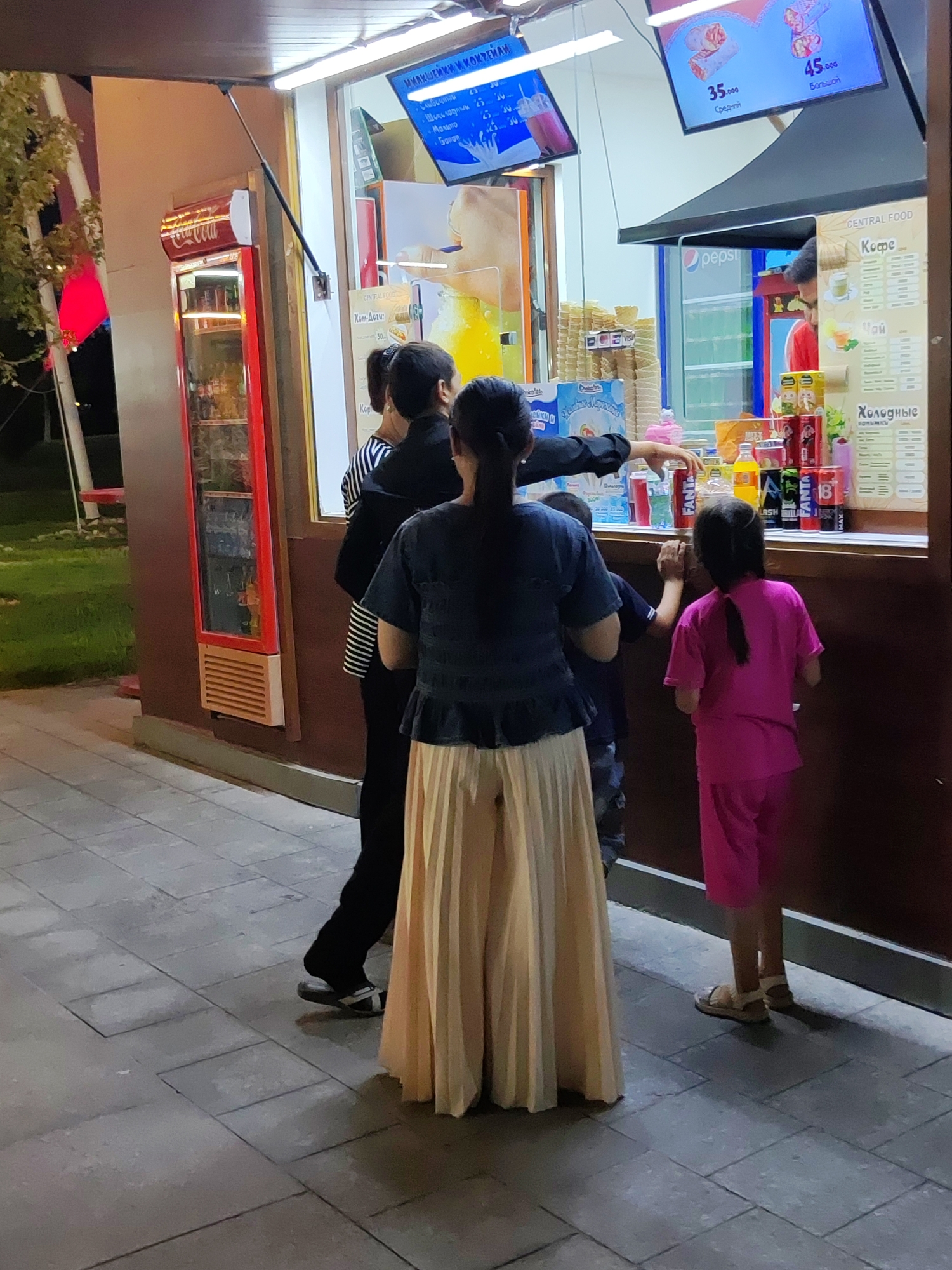 The image depicts a street food stall in Tashkent, Uzbekistan, at night. A family, including two adults and two children, are gathered at the counter, appearing to make a purchase. The stall sells beverages and snacks, with various drinks displayed in a refrigerator and on shelves. The lighting is artificial, illuminating the scene and highlighting the products. The ground is paved with gray tiles. The overall atmosphere suggests a casual evening outing.