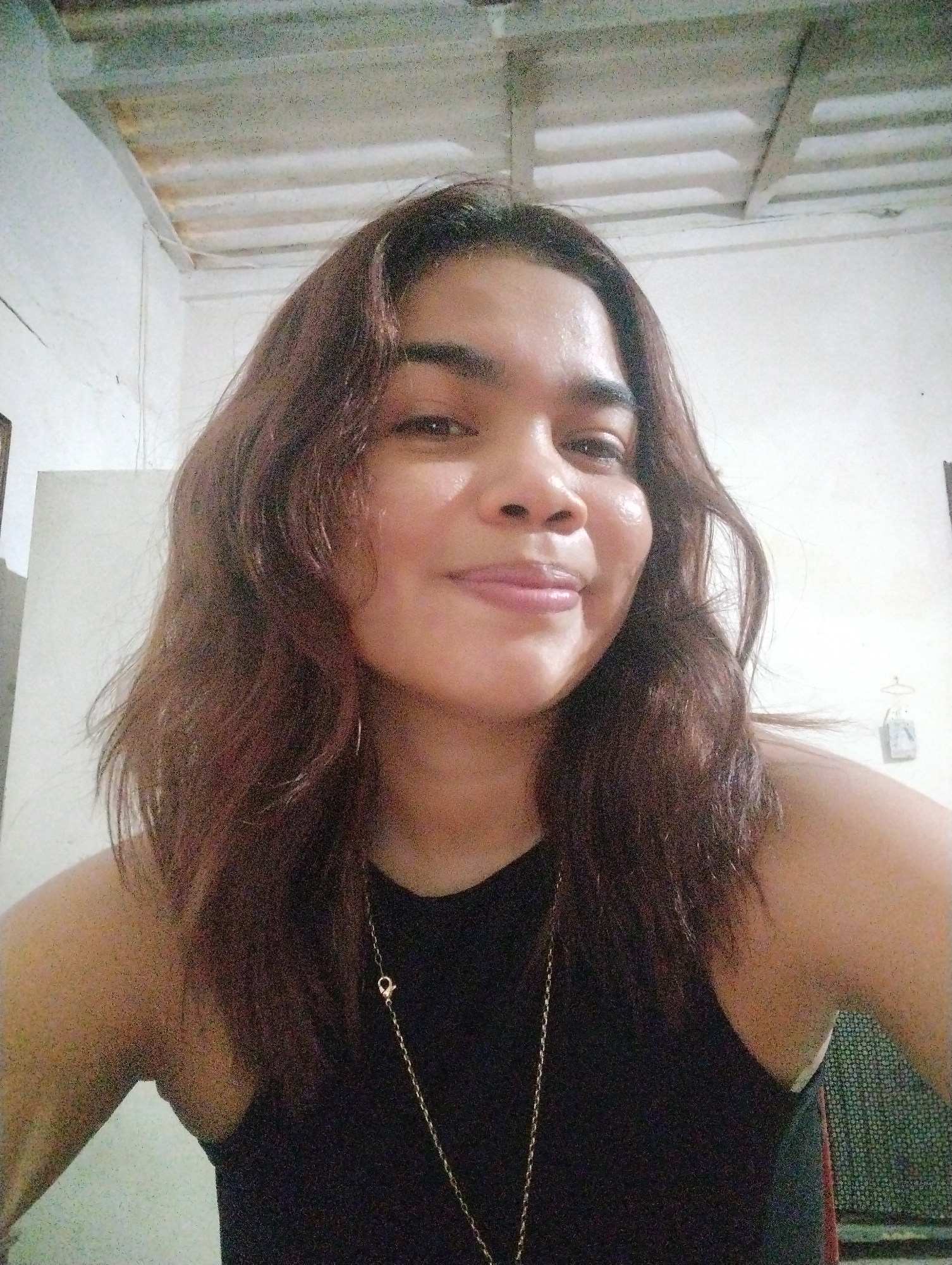A medium shot shows a young woman from the chest up, looking directly at the camera with a slight smile. She has dark brown, wavy hair that falls to her shoulders and is wearing a black tank top with a thin gold necklace. The background is a plain, white wall with a textured ceiling visible at the top. The lighting appears to be natural and diffused, casting a gentle glow on her face, which appears slightly oily. The overall impression is casual and informal, likely taken indoors in a domestic setting within Punta de Piedras, Venezuela.