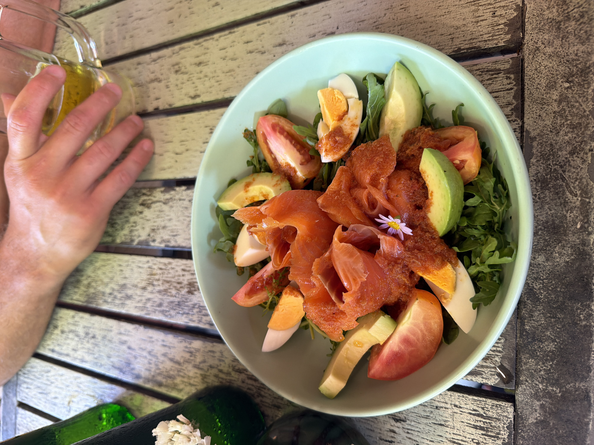 A high-angle, medium shot captures a salad in a light green bowl, placed on a weathered wooden table. The salad features smoked salmon, sliced hard-boiled eggs, avocado, and tomato wedges, atop a bed of green leaves. A small purple flower garnishes the salmon. To the left of the bowl are two green bottles, one with a white paper label. In the upper right, a person's hand is seen holding a glass mug filled with a golden liquid, likely beer, with a portion of their arm visible. The table's surface is made of wooden planks with visible grain and texture, suggesting an outdoor or rustic setting. The overall lighting is bright, casting shadows. This scene is taking place at a restaurant in Karon, Thailand.