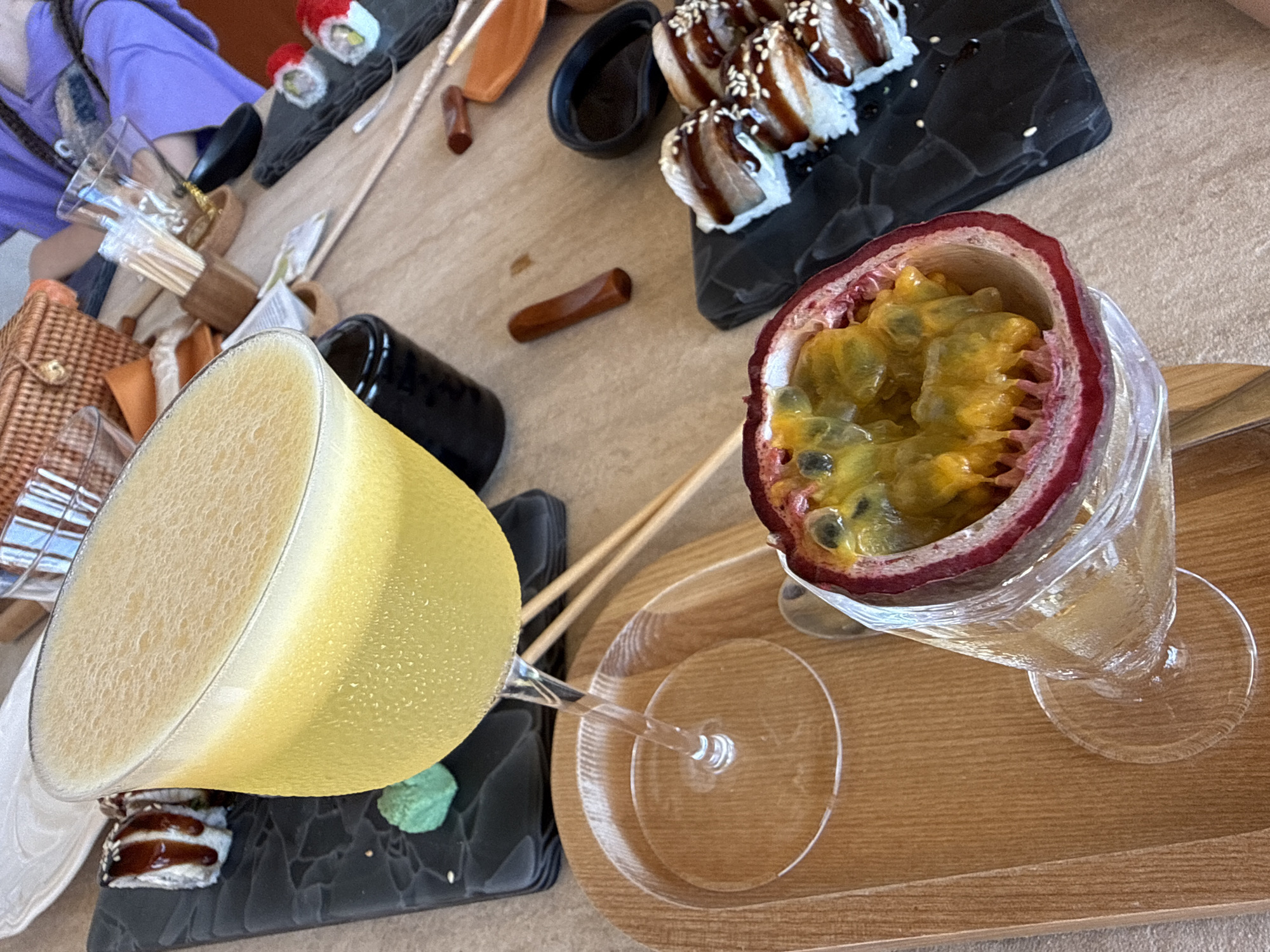 The image shows a close-up view of a table with food and drinks. In the foreground, a halved passion fruit is presented in a glass. Behind it, a yellow cocktail in a coupe glass is visible, with a frothy top. To the left and right of the cocktail, plates of sushi are arranged. The table setting includes wooden chopsticks, a small dish of green paste (likely wasabi), and a soy sauce dish. In the background, a wicker basket and parts of people are visible, suggesting a casual dining setting. The lighting suggests it is daytime. The image does not contain any information about the location being Alushta, Ukraine, or any other specific city.