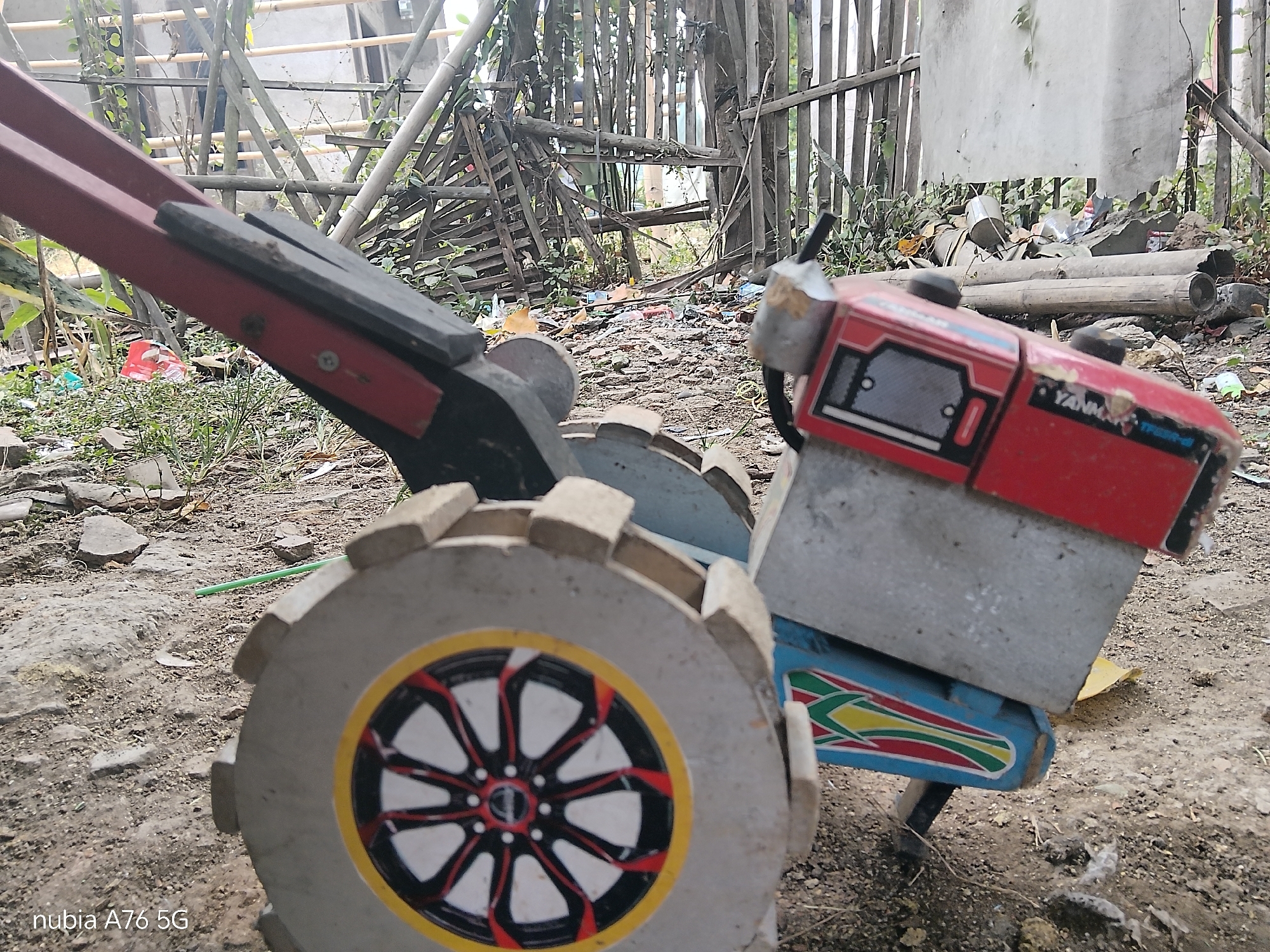 The image shows a close-up, ground-level view of a red and gray Yanmar tilling machine, possibly a rotary tiller, parked on a dirt ground covered with small stones and debris. The machine has a large, circular tire with a decorative pattern featuring red, white, and black spokes within a yellow rim. A portion of the red engine housing is visible, with the "YANMAR" logo clearly displayed in white text. The machine is positioned in an outdoor environment, with a rustic fence made of vertical wooden poles and horizontal bamboo supports in the background. Vegetation, including tall grass and leafy plants, is growing around the machine and the fence. Scattered debris, including plastic bags and what appears to be discarded building materials, litters the ground. The overall scene suggests a rural or semi-rural setting in the city of Binuang, Indonesia, possibly a storage area or a worksite where the tilling machine is kept. There are no people visible in the image.