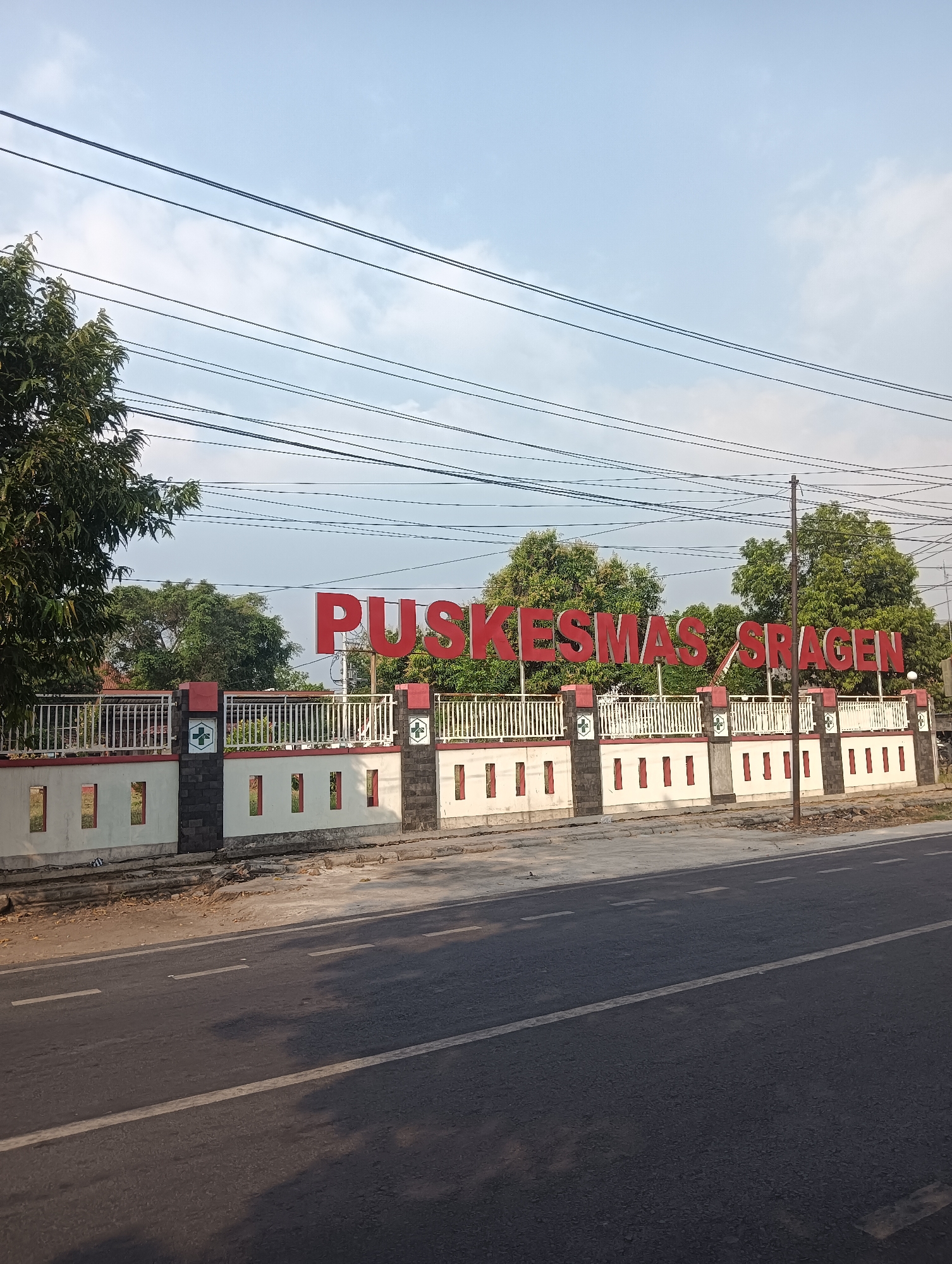 The image shows a street scene in Sragen, Indonesia, in front of a building identified by a large red sign that reads "PUSKESMAS SRAGEN." The sign is mounted on a low white wall with decorative red and black stone pillars. Behind the wall, trees and some buildings are visible. The foreground consists of a paved road with white lane markings. The sky is partly cloudy. There are no people visible in the image. The overall environment appears to be an outdoor urban or semi-urban setting.