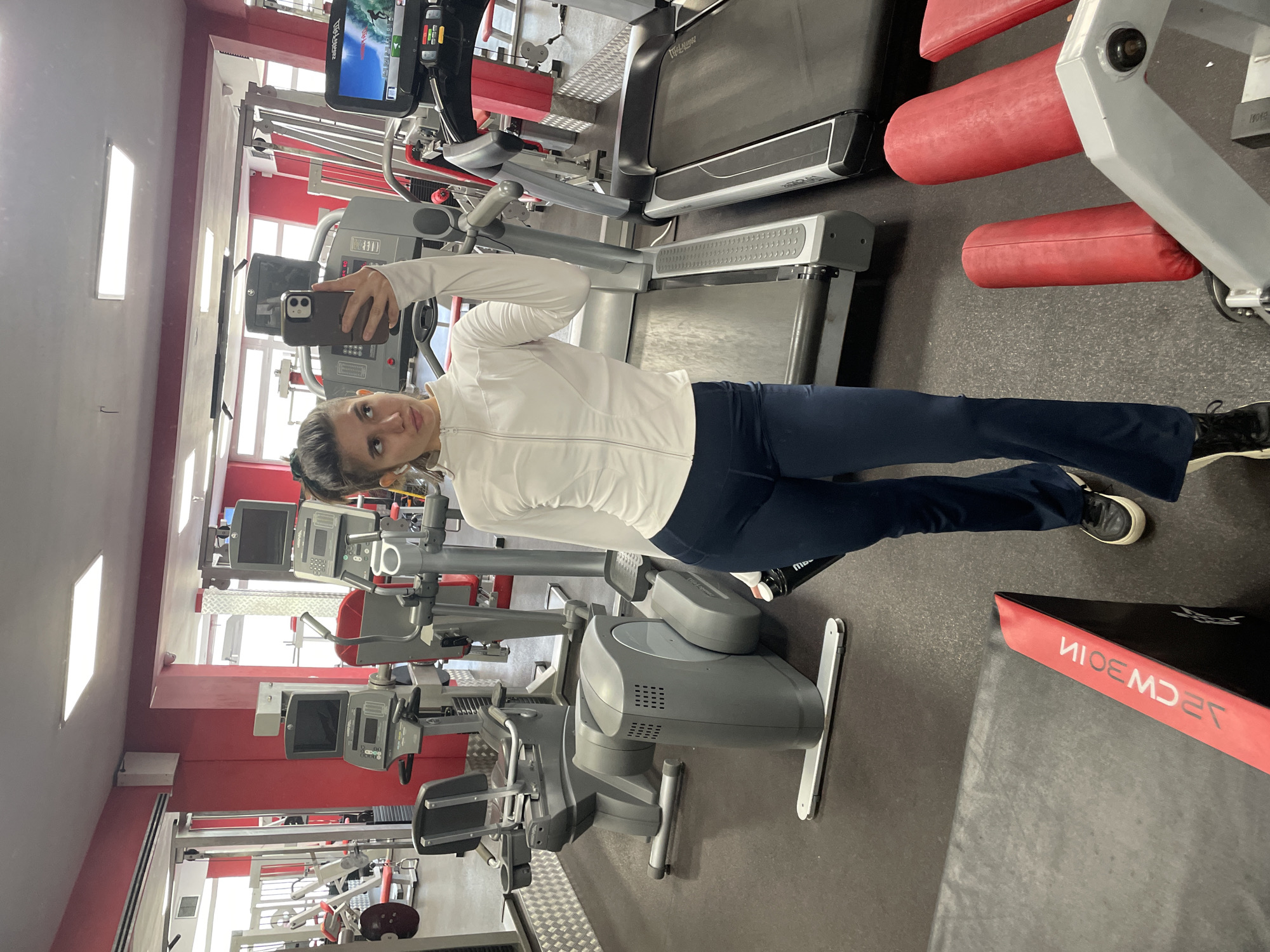 The image is taken at a gym in Sofia, Bulgaria, indicated by the Cyrillic writing on the equipment in the foreground. A woman in workout attire, consisting of a white zip-up jacket and dark blue pants, is taking a selfie in the gym. She is standing in the middle of the gym floor, surrounded by various pieces of exercise equipment including a treadmill, stationary bikes, and weight machines. The gym has red and gray decor.